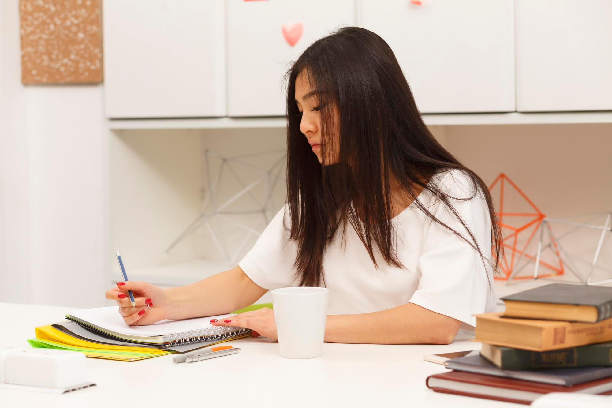 A woman seated at a desk, writing in a notebook, with a cup of coffee beside her.