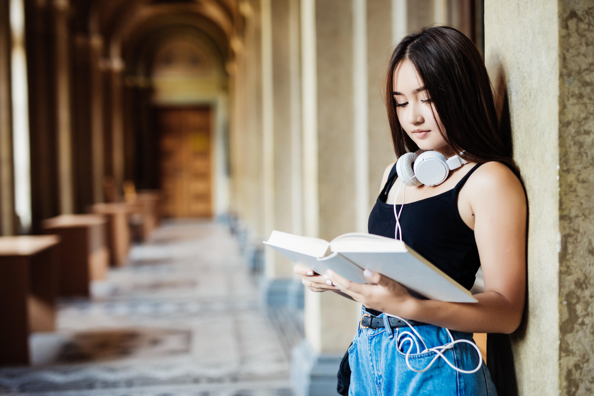 Asian woman reading a book in a corridor, illuminated by natural light, creating a serene and focused environment.