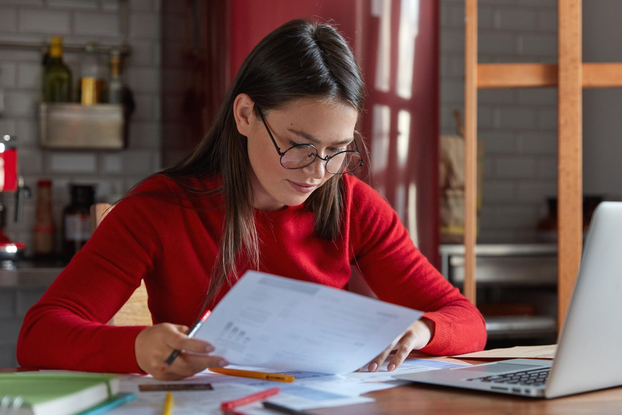 A woman with glasses examines documents on her laptop, concentrating on her tasks in a bright workspace.