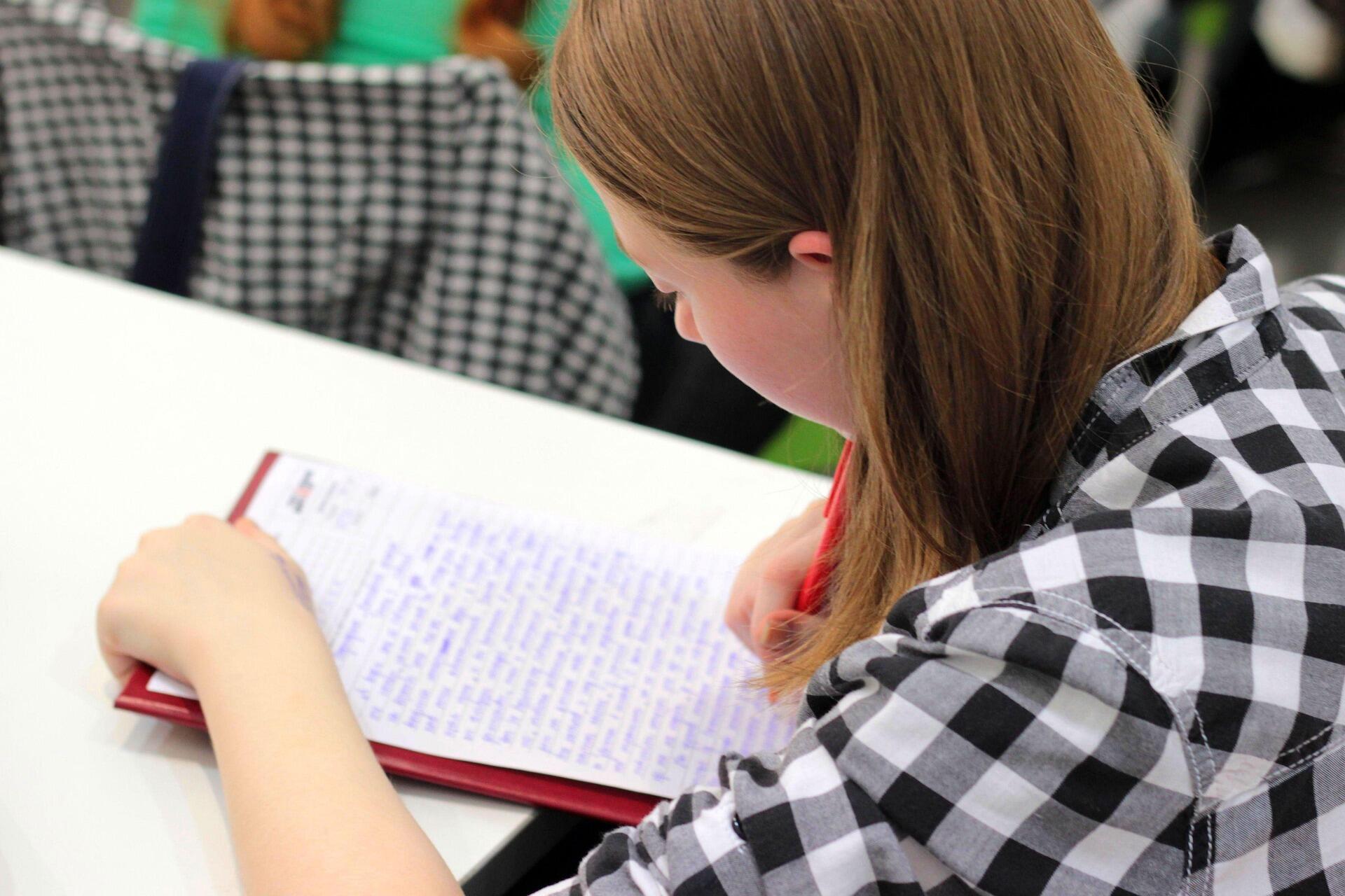 A young girl sits at a desk, writing intently in a notebook with a pencil in her hand.