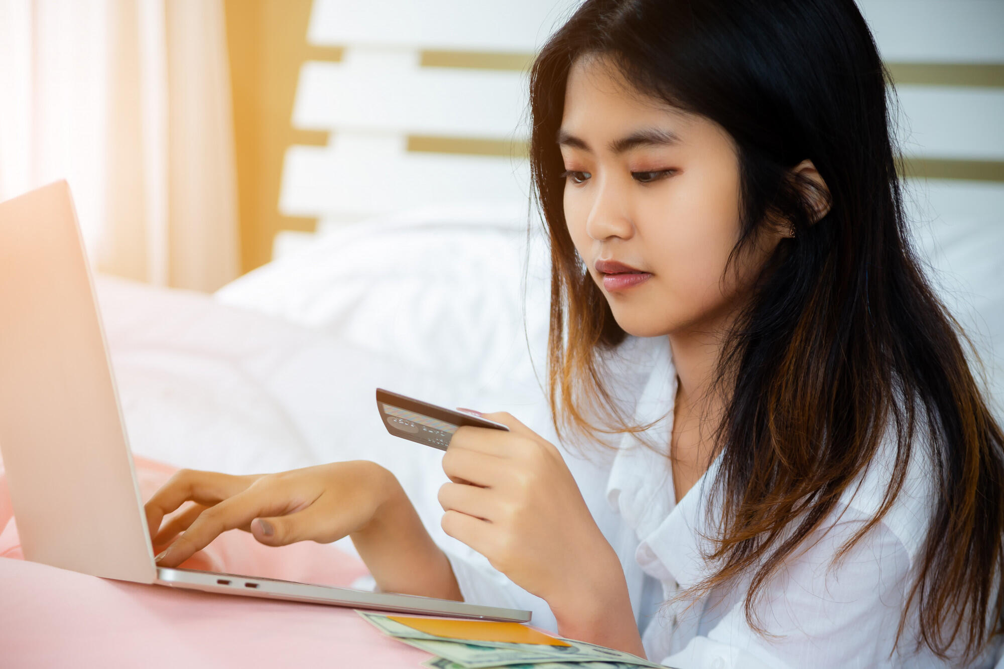 An Asian woman in bed, engaged with her laptop and a credit card, indicating online transactions or purchases.
