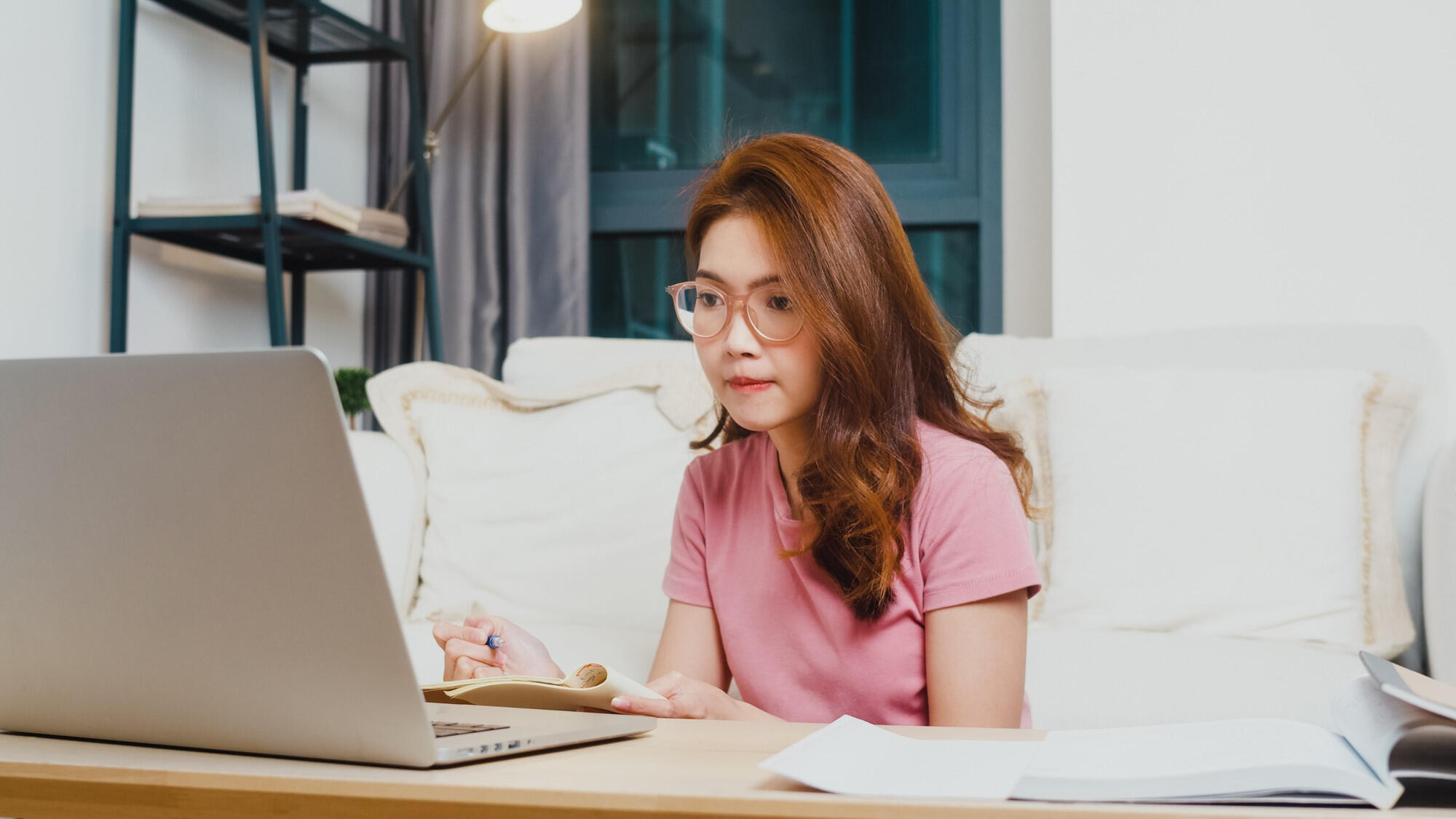 A woman wearing glasses sits at a table, focused on her laptop while working or studying.
