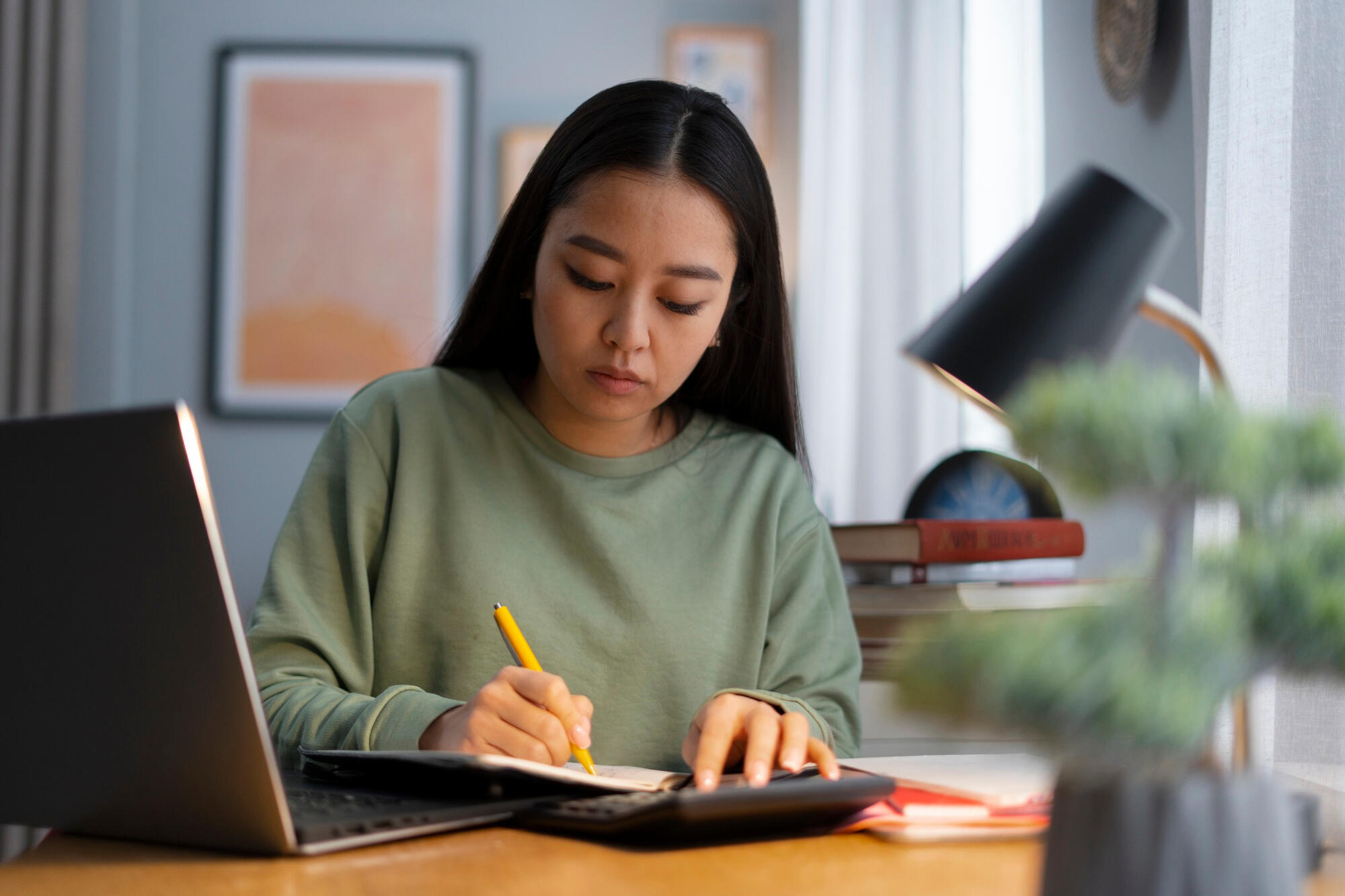 A woman focused on her laptop, seated at a desk with a notepad and pen beside her.