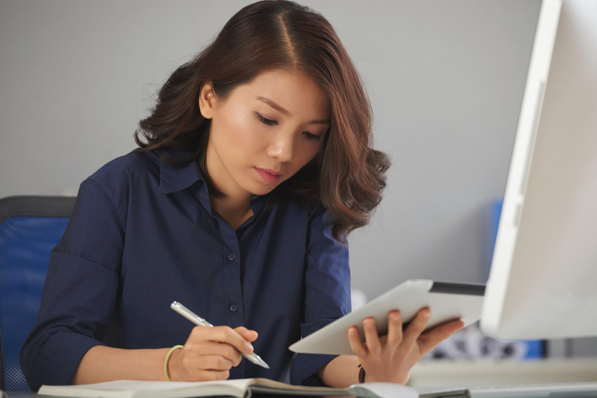 A woman in a blue shirt writes on a tablet, focused on her task in a well-lit environment.