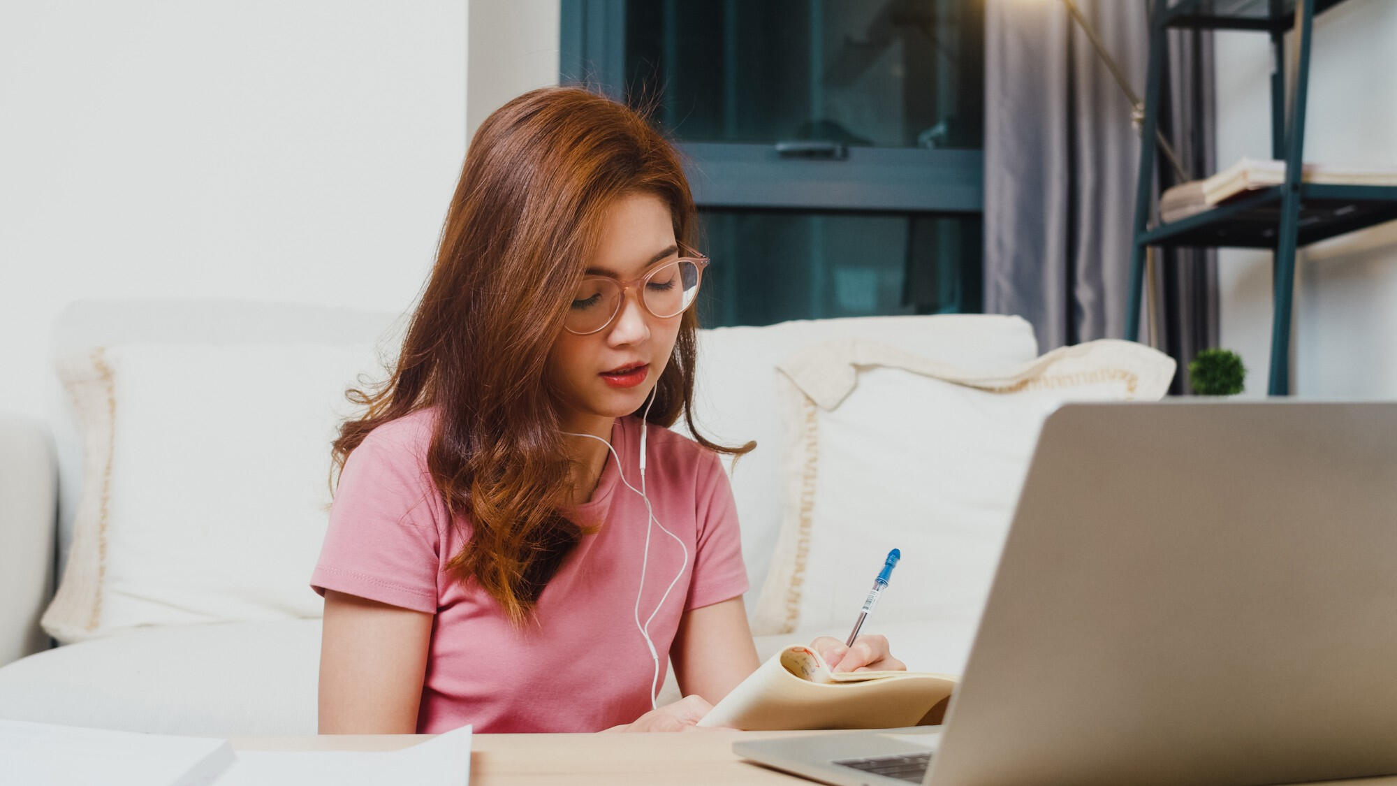 A woman in a pink shirt sits at a table, focused on her laptop while working or studying.
