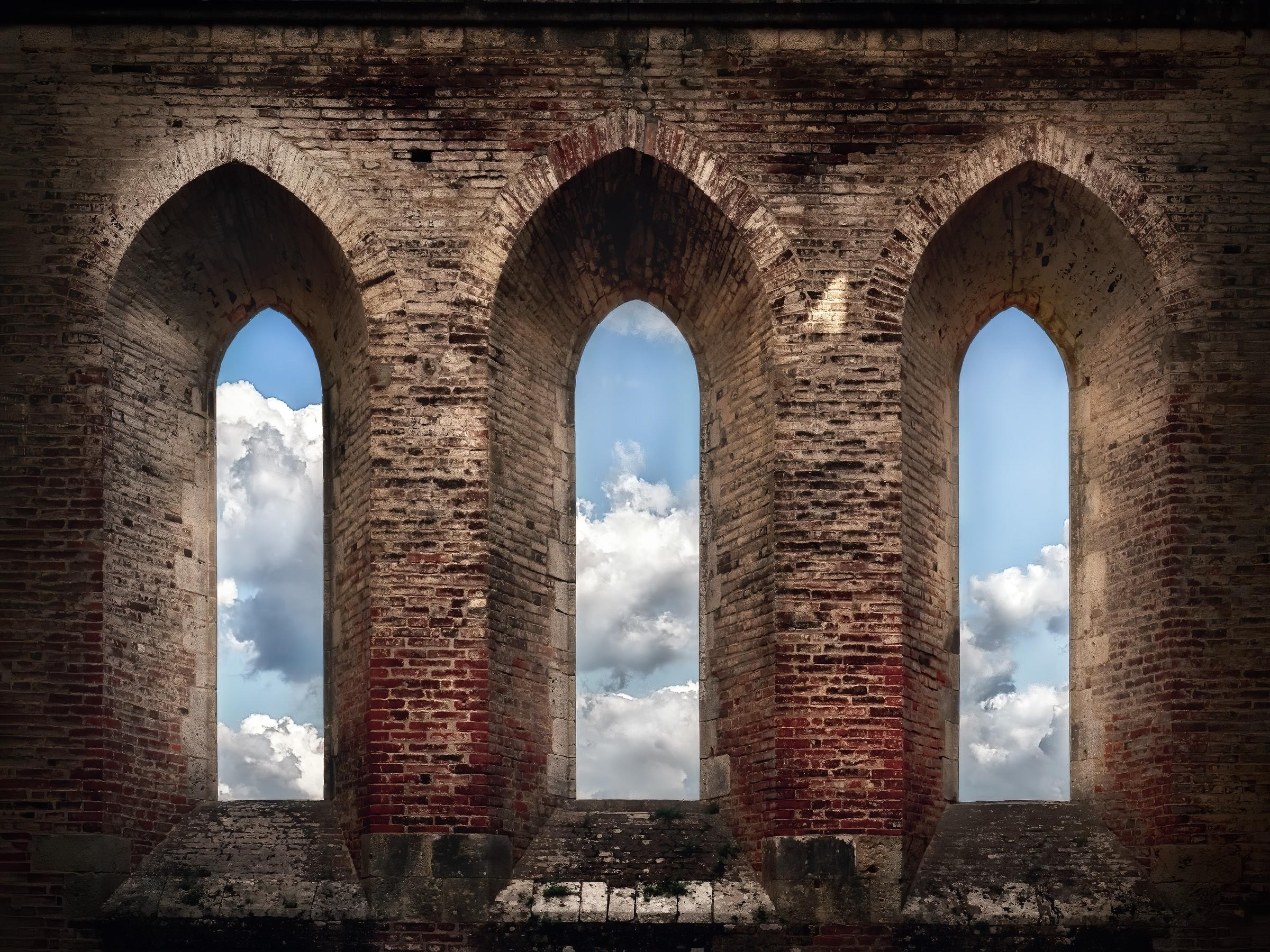 Een kasteelmuur met drie ramen erin, waardoorheen je wolken en lucht ziet
