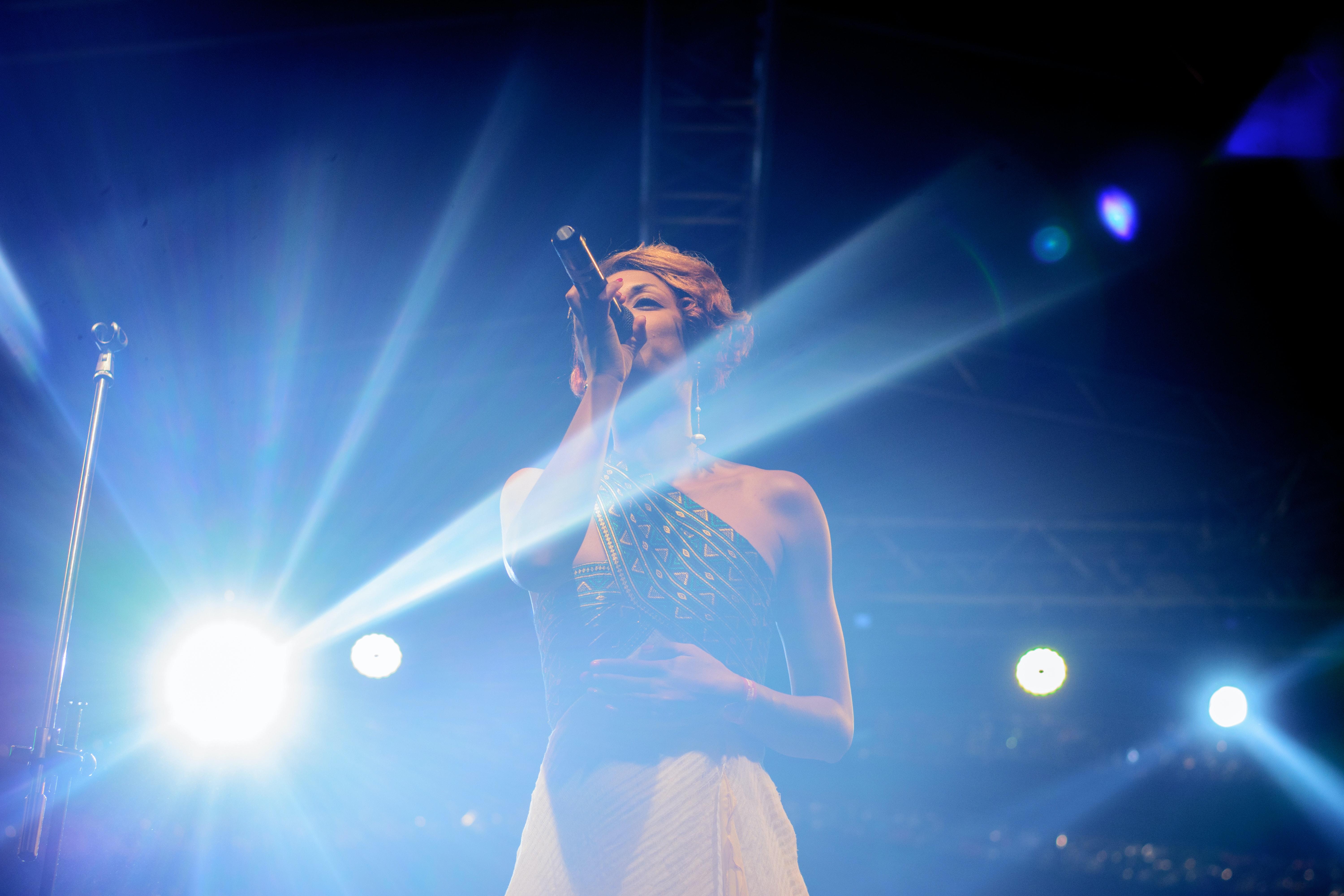 Vrouw zingt op podium met microfoon en lampen.