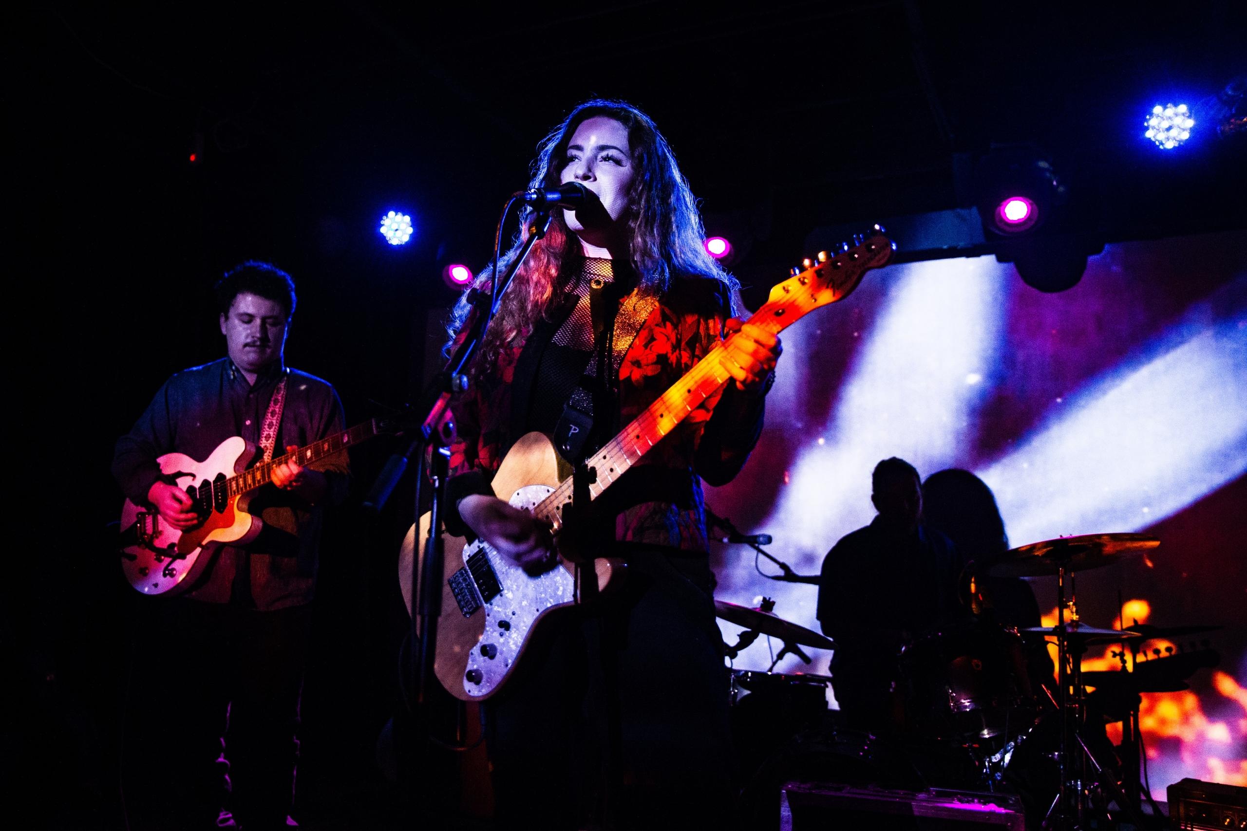 Vrouw zingt in een microfoon met een gitaar, met band op de achtergrond.