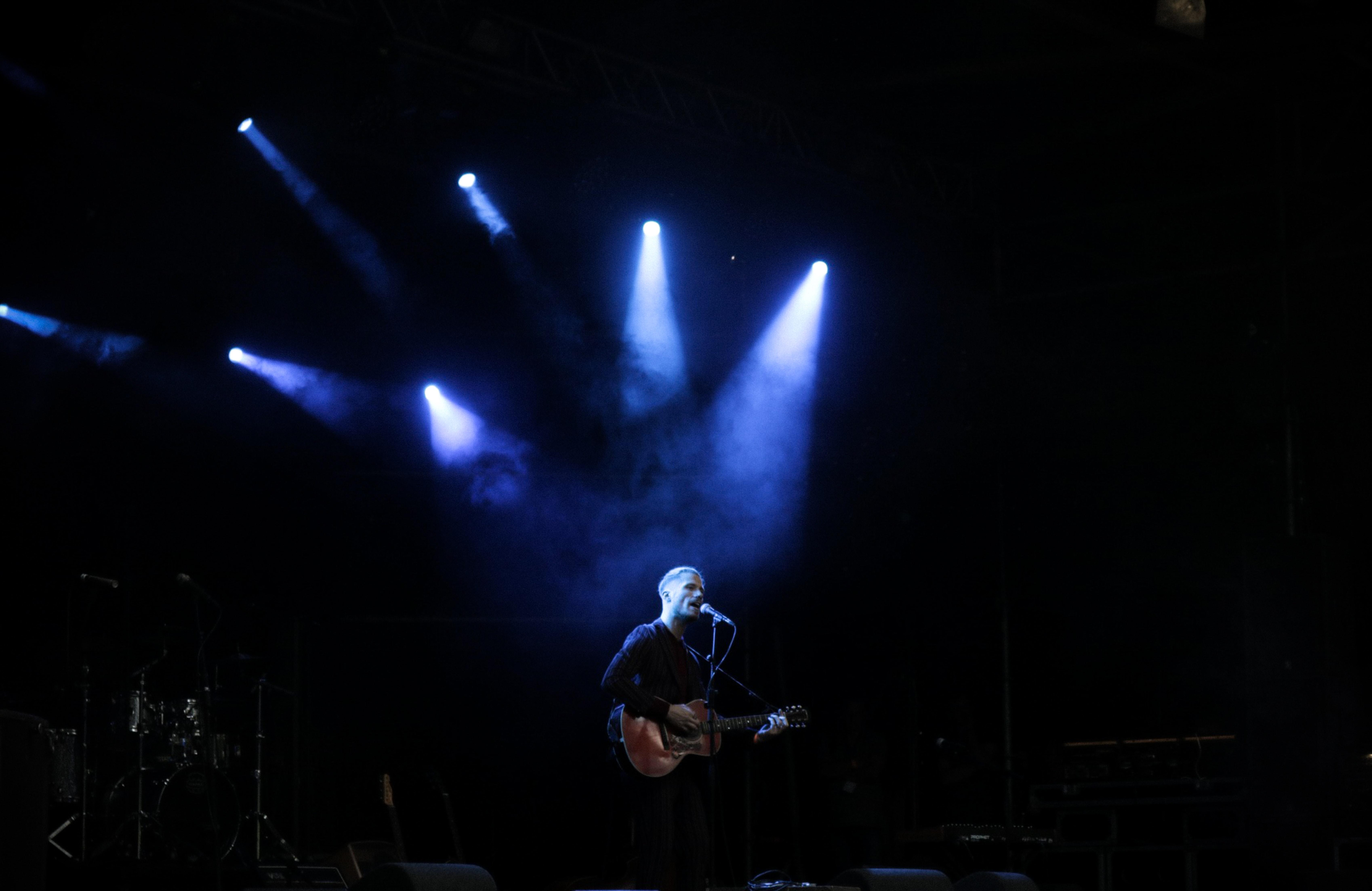Man zingt op podium met gitaar in een microfoon, met lampen die op hem gericht zijn.