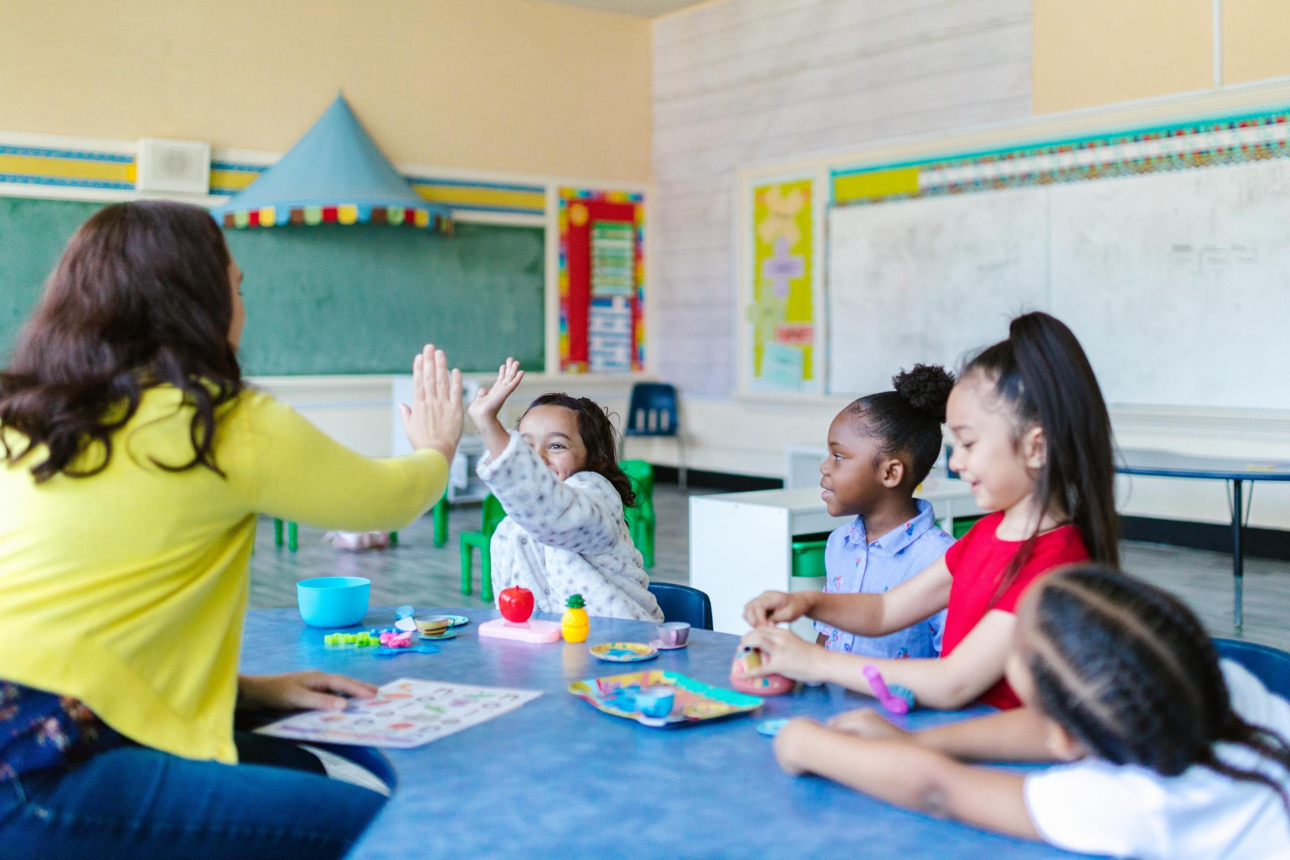 Kinderen zitten aan een tafel in een klaslokaal met hun leraar te kleuren en te spelen met objecten.
