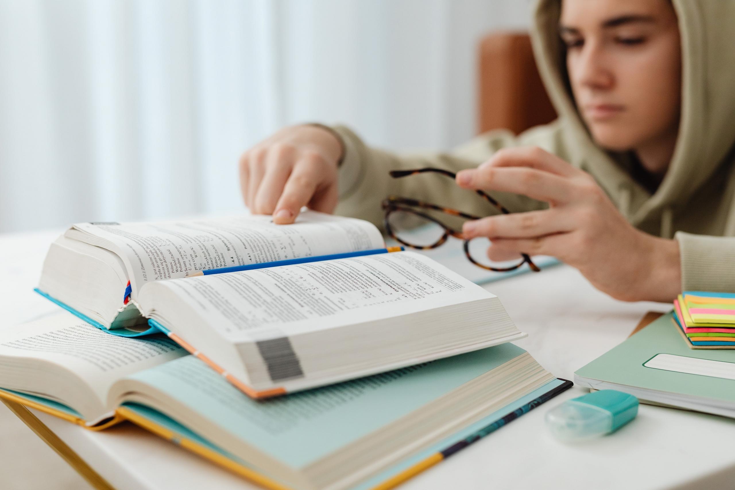 Jongen met capuchon zit aan tafel met boeken voor zich en een bril in zijn hand