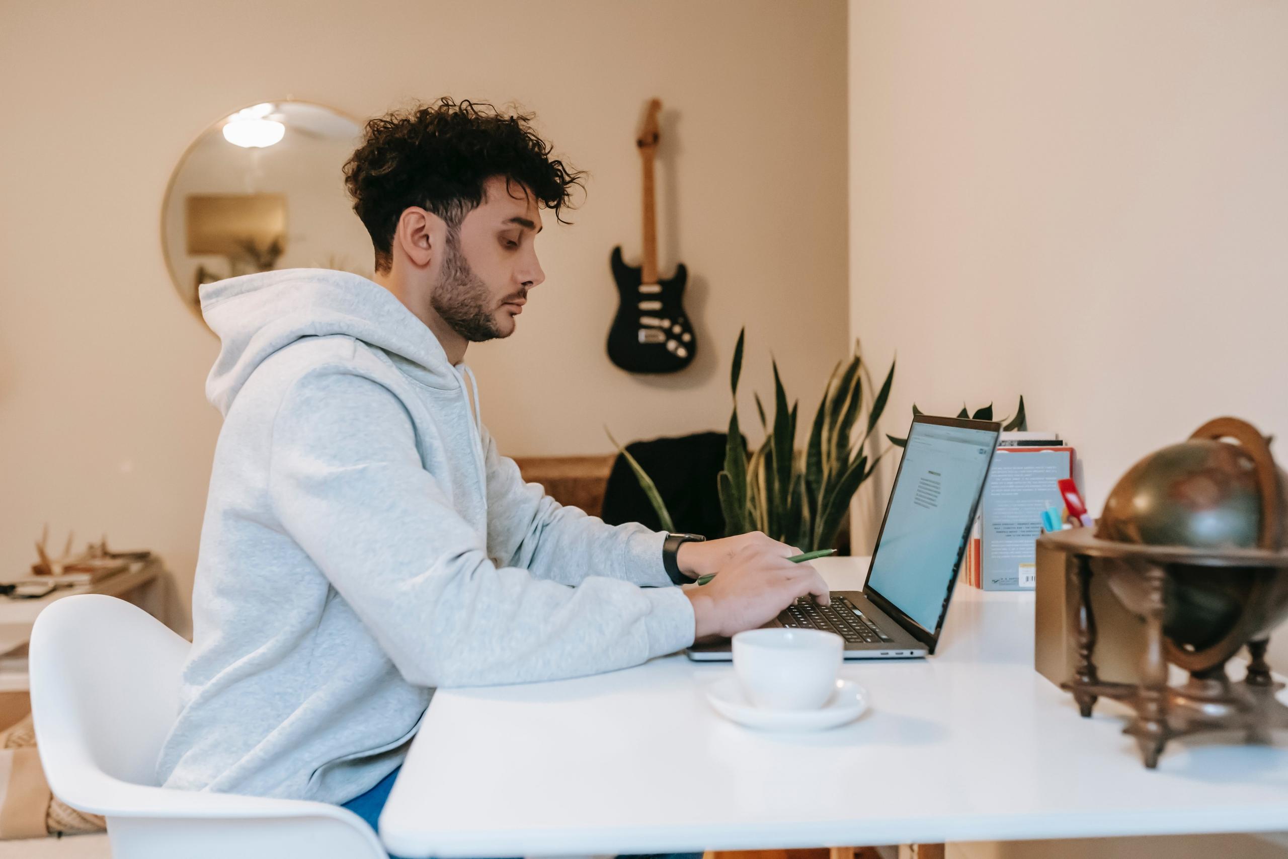 Een man die aan een tafel zit en werkt op een laptop, met een elektrische gitaar aan de muur.