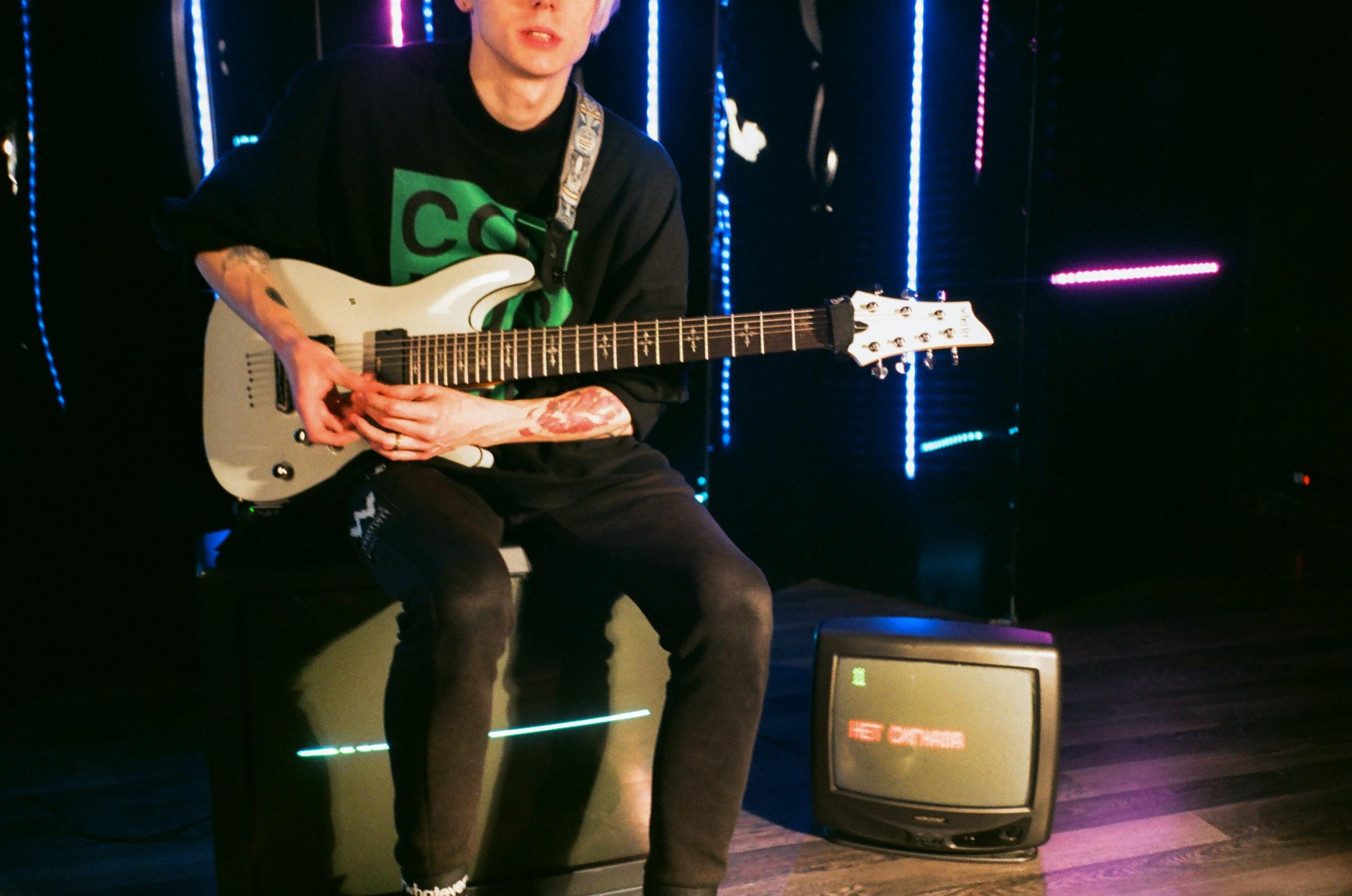 Een gitarist zit voor kleurrijke lichten met een televisie op de voorgrond.