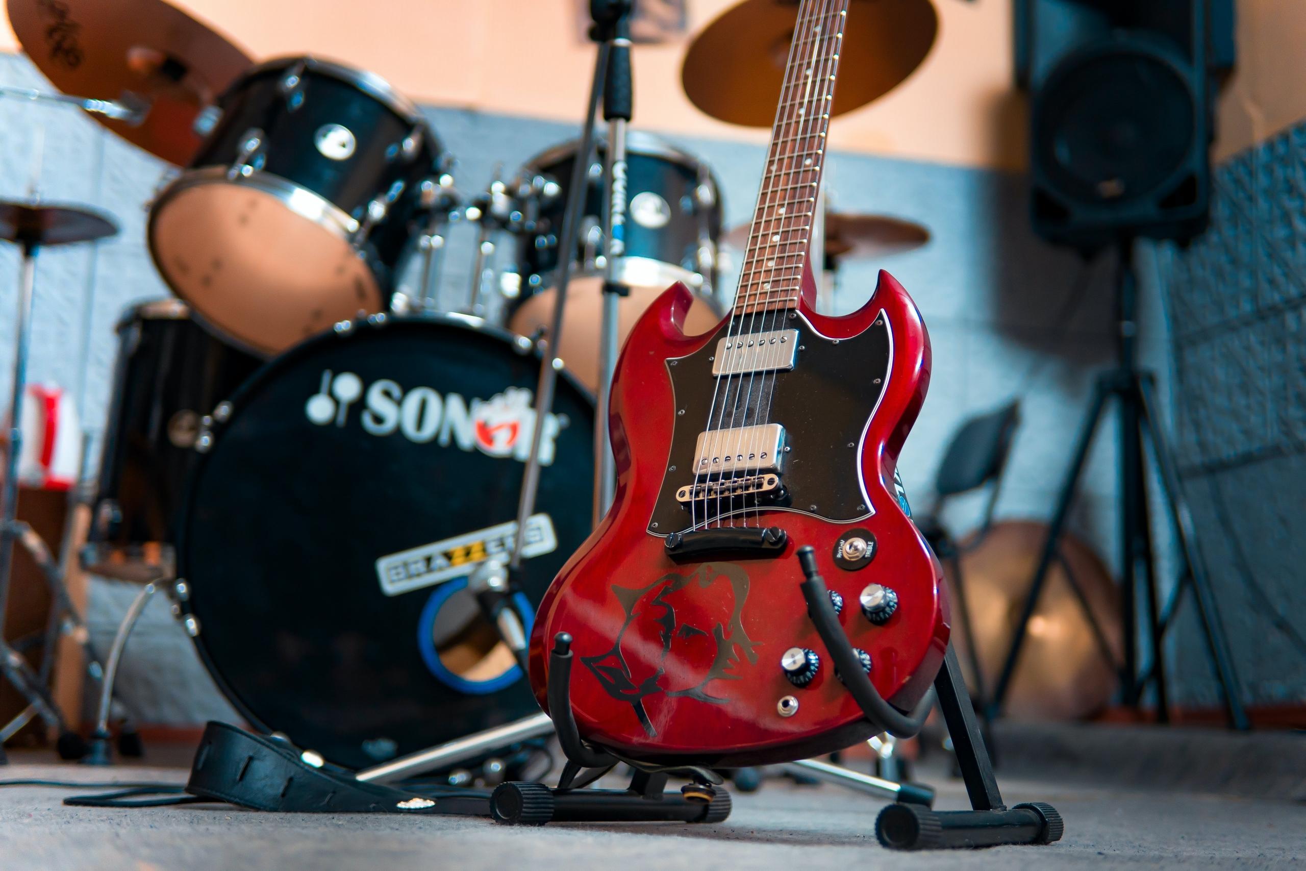 Een rode elektrische gitaar rustend op een standaard met een drumstel op de achtergrond.