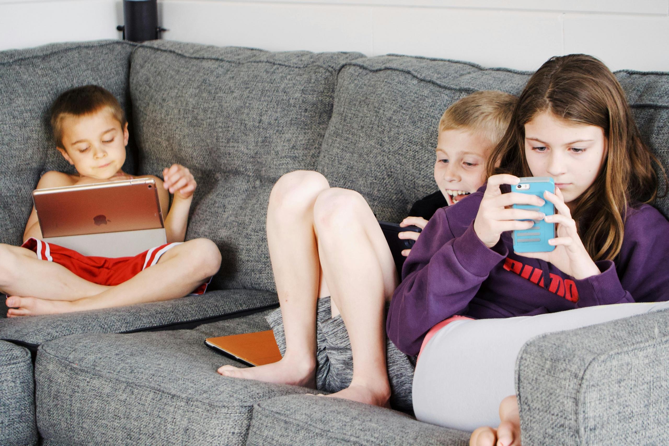 Drie kinderen zitten op een bank, elk met een eigen apparaat; een tablet en een smartphone op de bank.