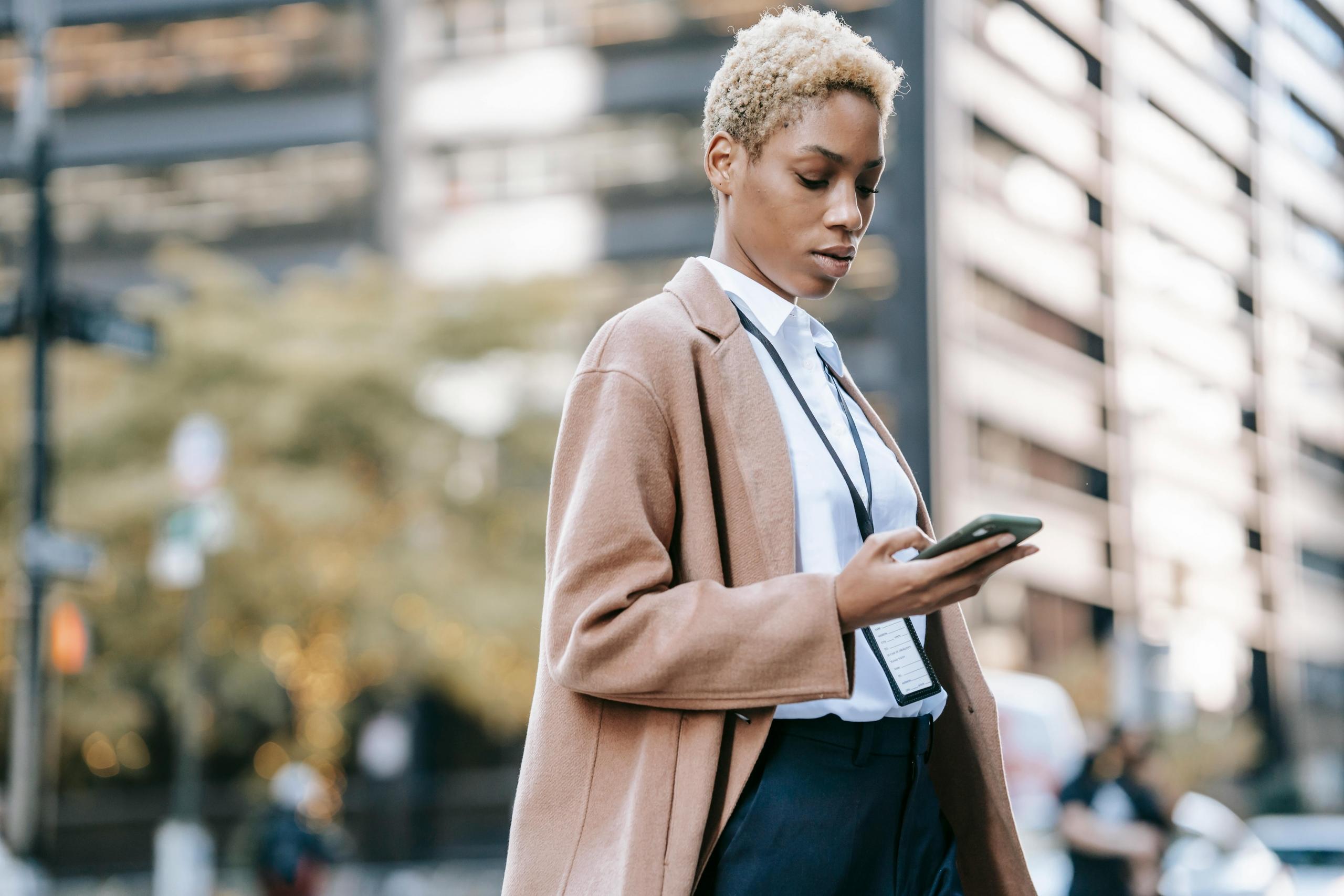 Een zakelijk geklede vrouw gebruikt haar smartphone op straat met een stedelijke achtergrond.