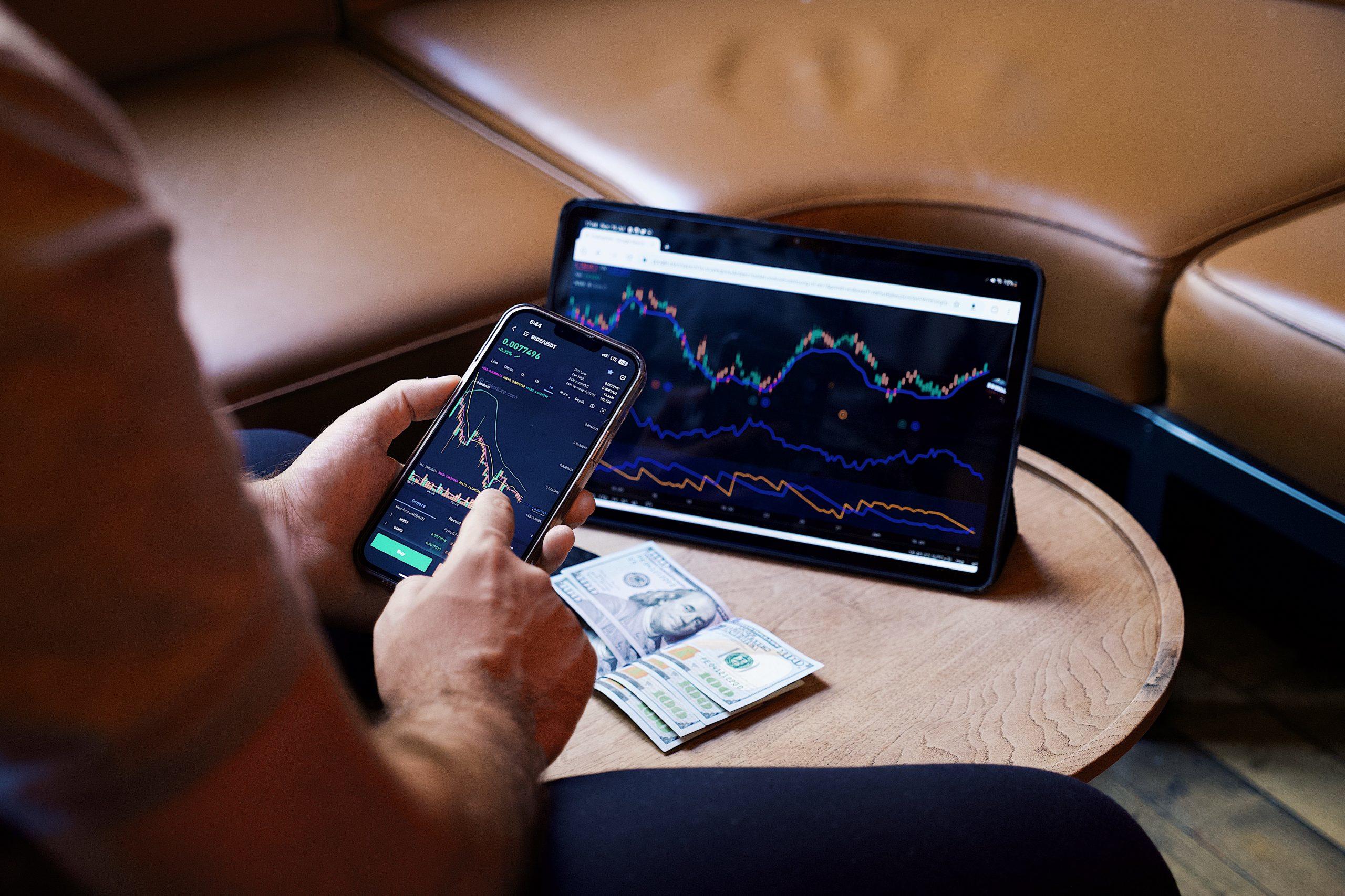 Een persoon gebruikt een smartphone en tablet tegelijkertijd voor financiële informatie met contant geld op tafel.