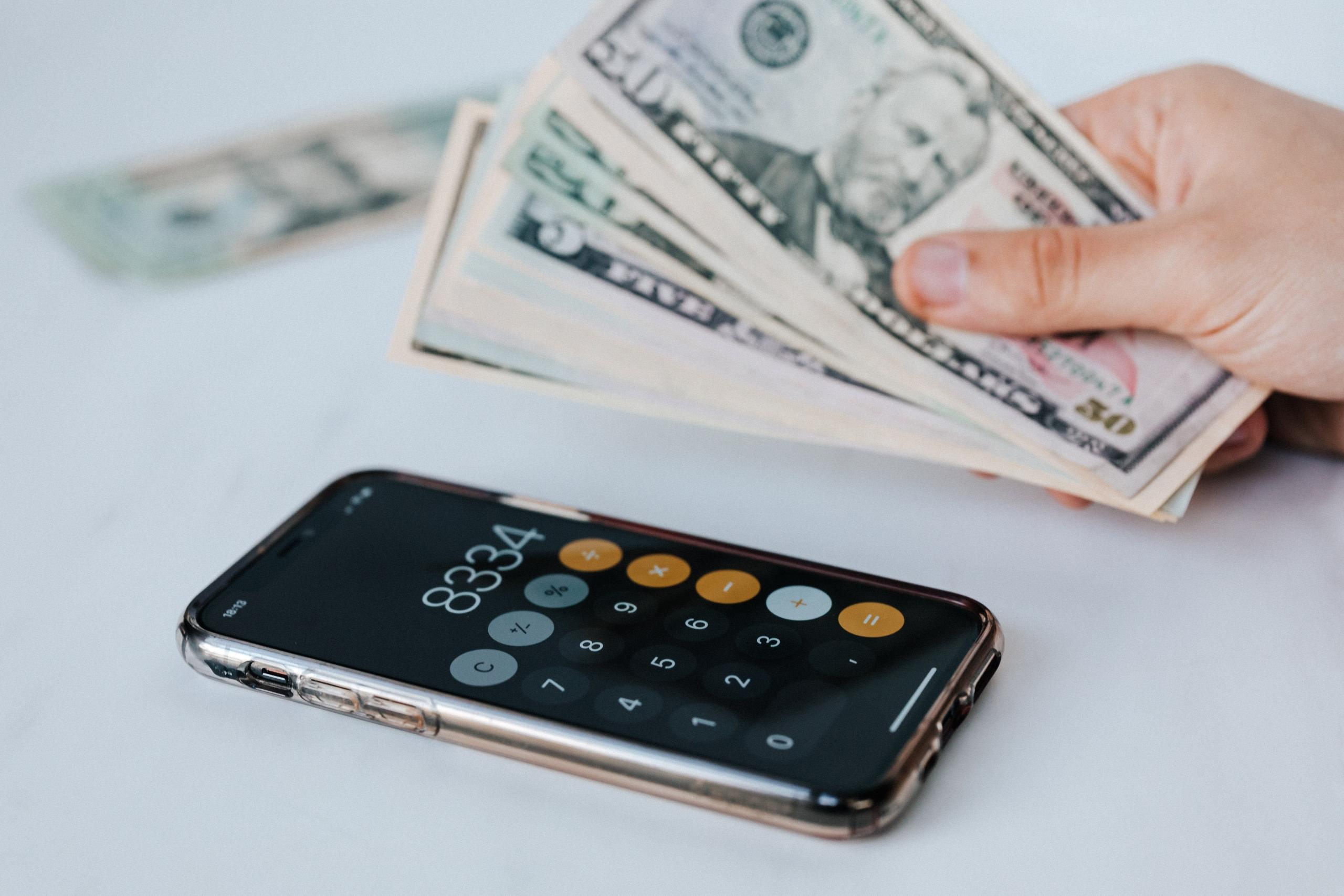 Een hand houdt Amerikaanse dollarbiljetten boven een smartphonecalculator.