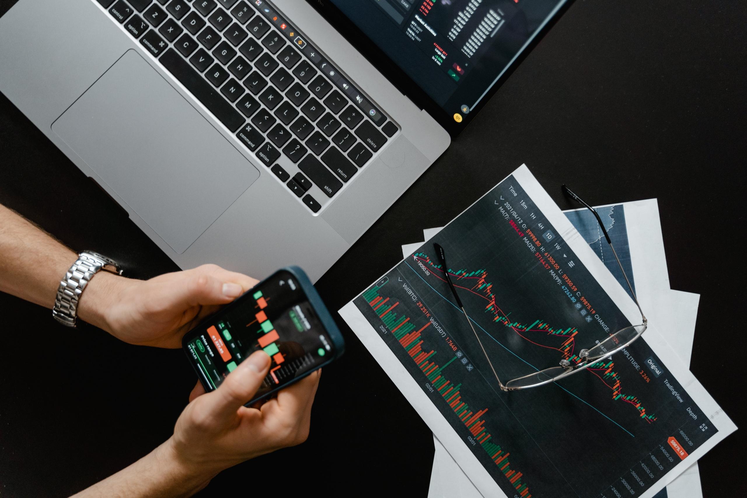 Een persoon gebruikt een smartphone boven een laptop met financiële grafieken op het scherm en papieren grafieken ernaast.