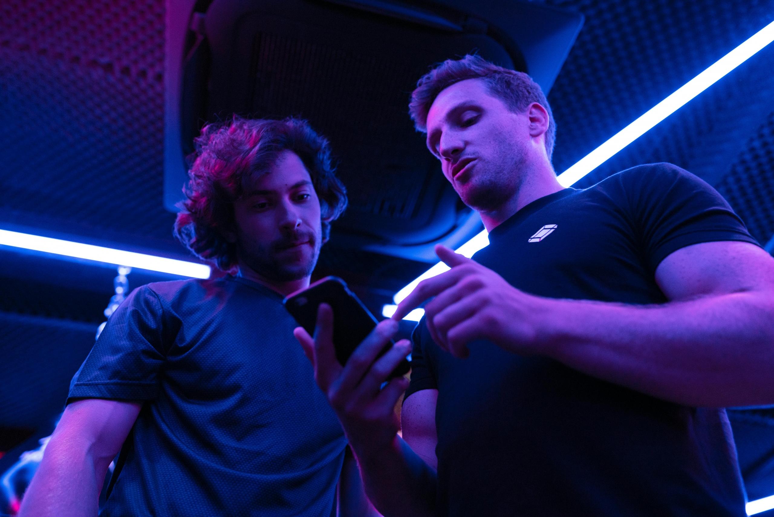 Twee mannen kijken samen naar een smartphone in een ruimte met blauwe verlichting.