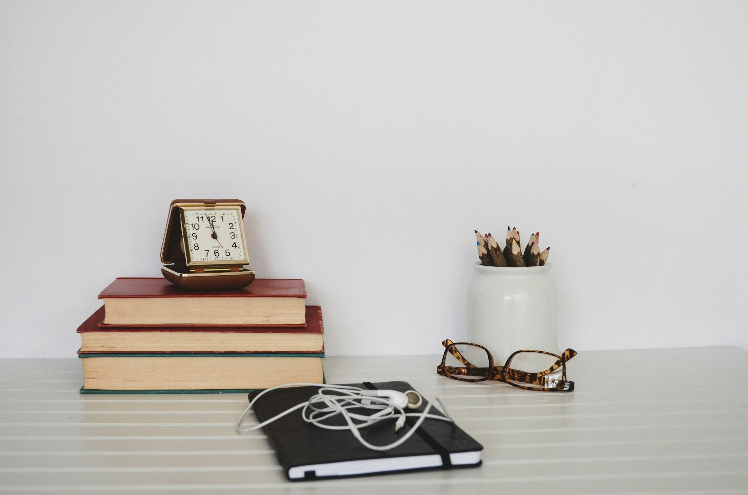 Klokje op stapel boeken, bril, potloden, oordopjes en schrift