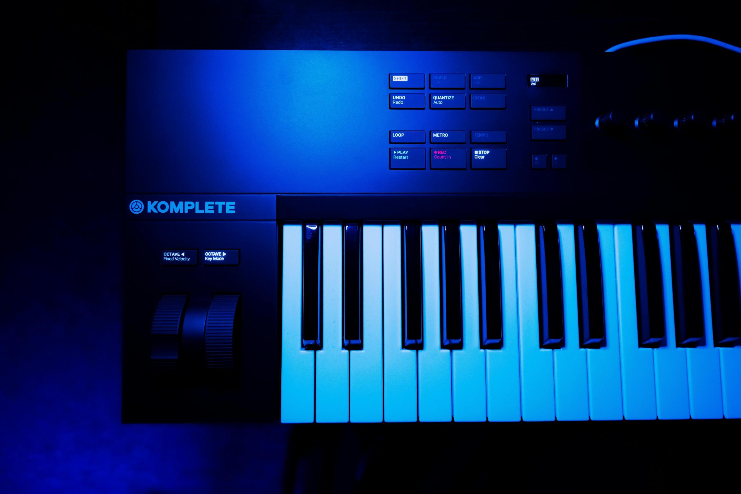 Een blauw verlicht MIDI-keyboard van het merk KOMPLETE. De verlichting benadrukt de zwarte en witte toetsen en de bedieningselementen aan de linkerkant.