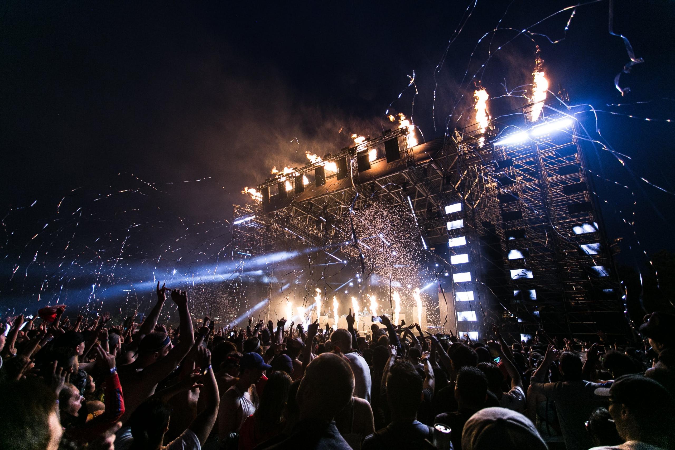 Een concert met een groot publiek dat naar een podium kijkt waar vuurwerk en lichteffecten te zien zijn.