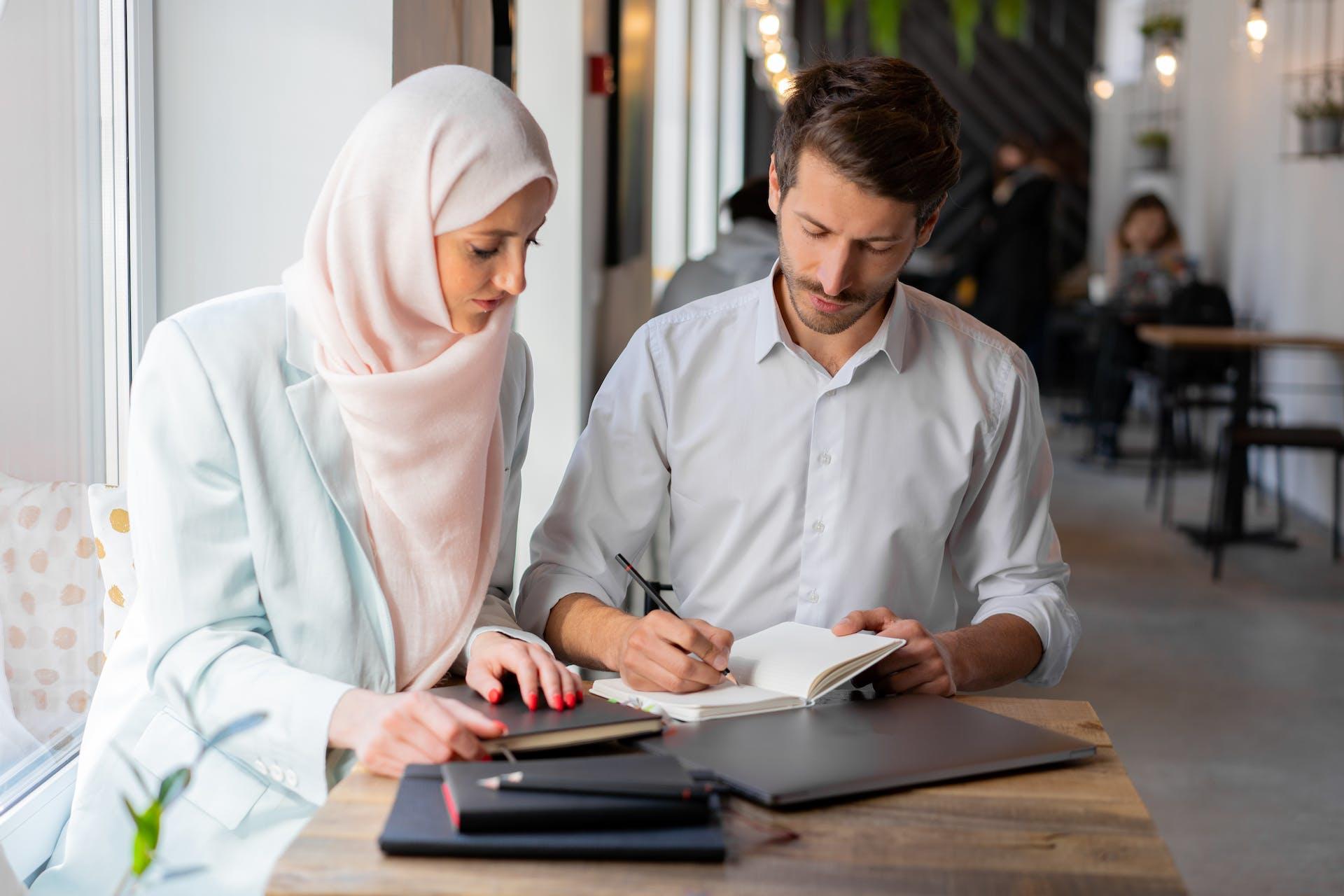 Vrouw met hoofddoek kijkt naar schrijvende man naast haar aan tafel
