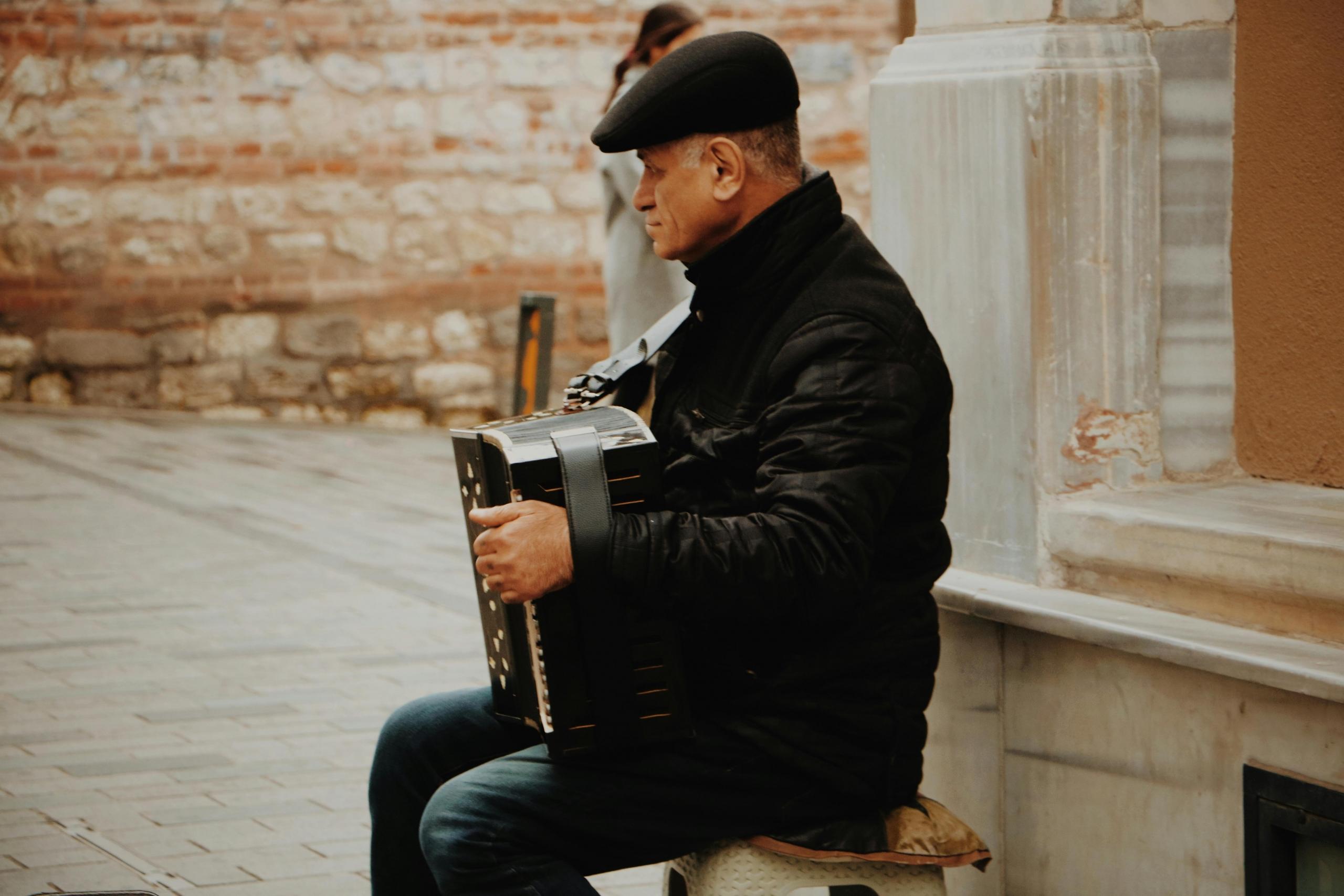 Een oudere man, zittend op een stoel buiten, gekleed in een zwart jack en een platte pet, spelend op een accordeon.
