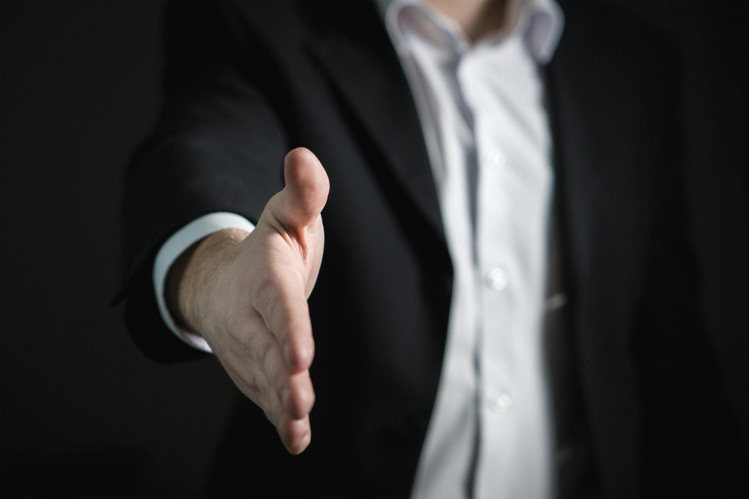 Een man in een donker pak reikt zijn hand uit alsof hij iemand begroet of een handdruk aanbiedt.