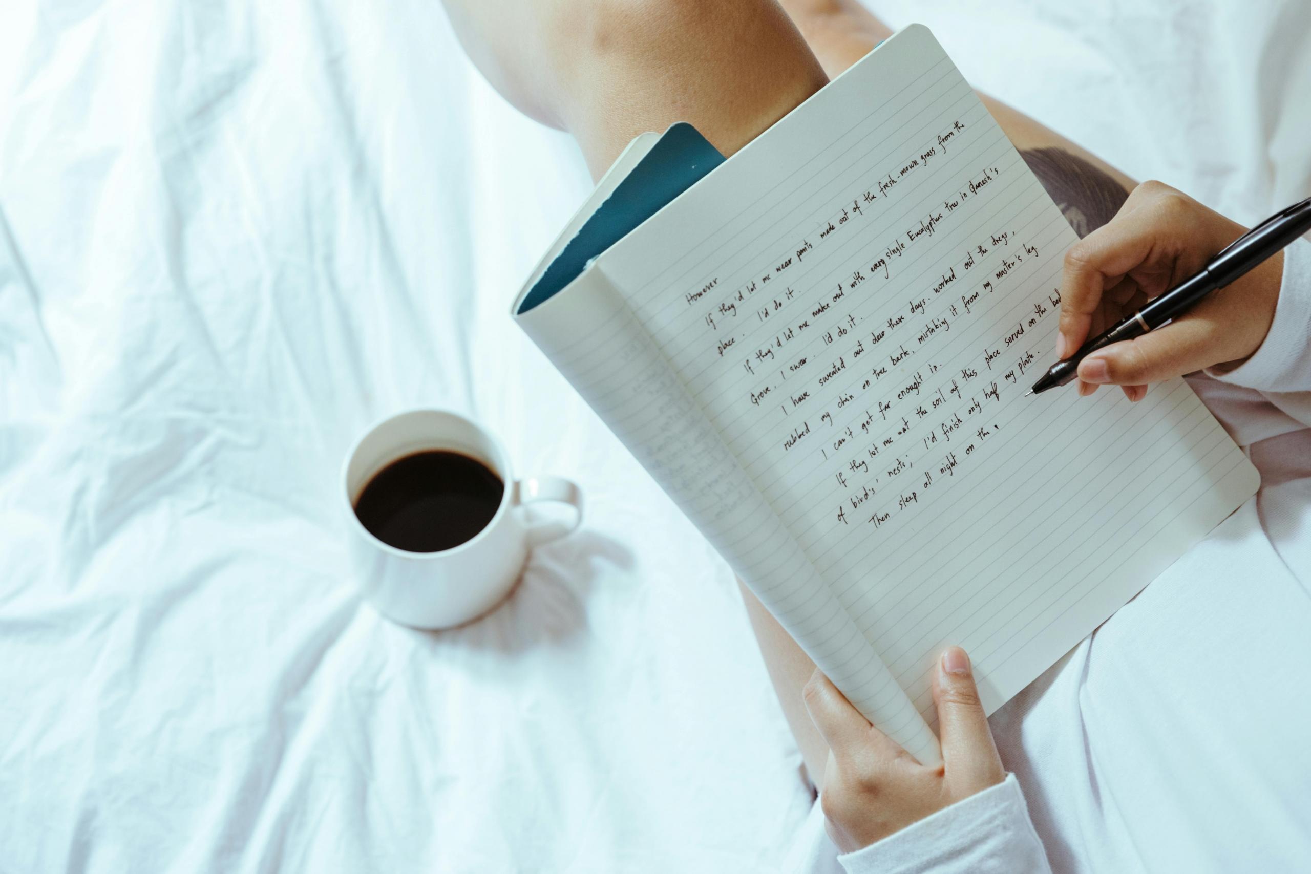 Iemand schrijft in een notitieboek dat op schoot ligt met een kop koffie ernaast.