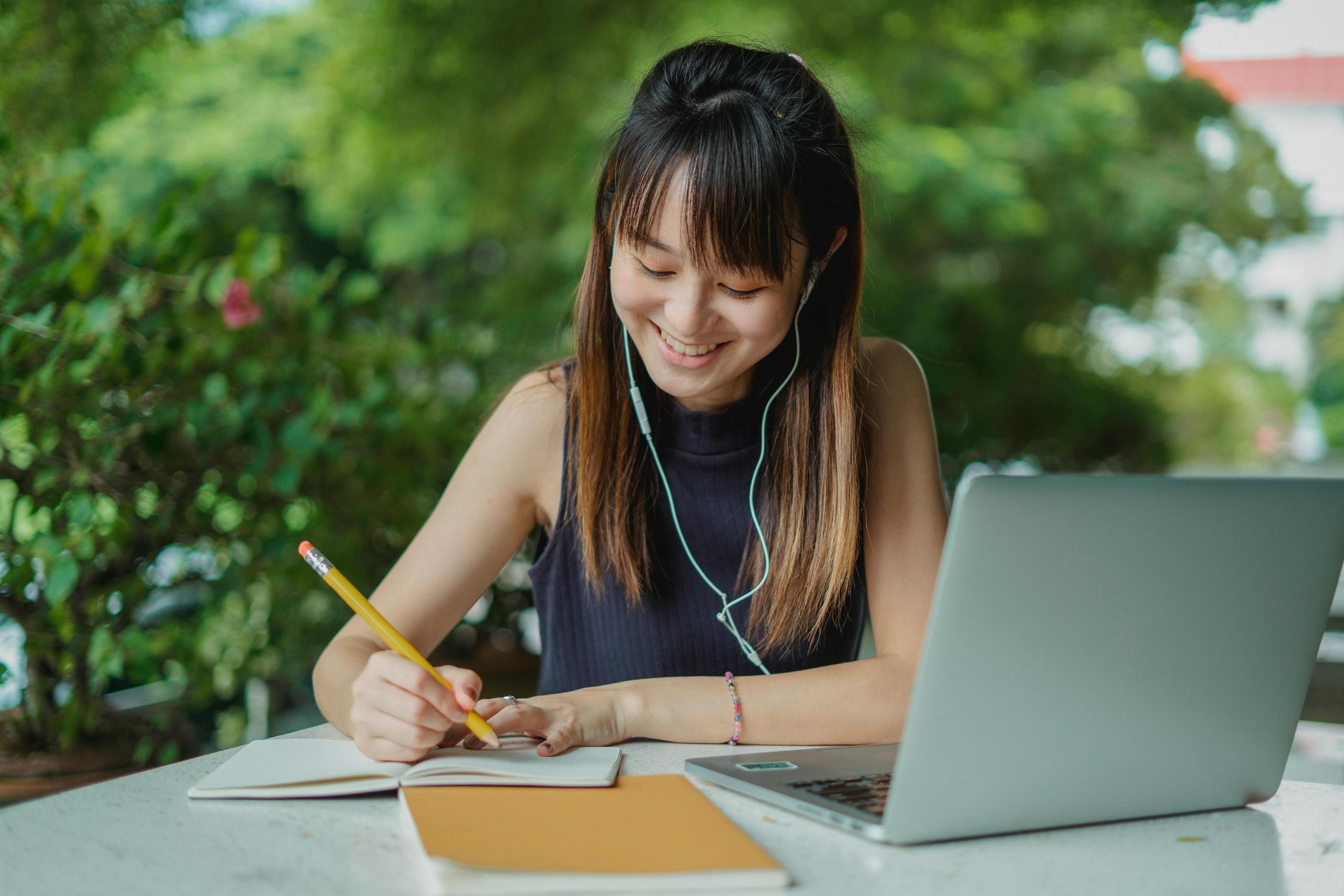 Een vrouw zit buiten te schrijven met een laptop en een boek op tafel terwijl ze oortelefoons draagt.