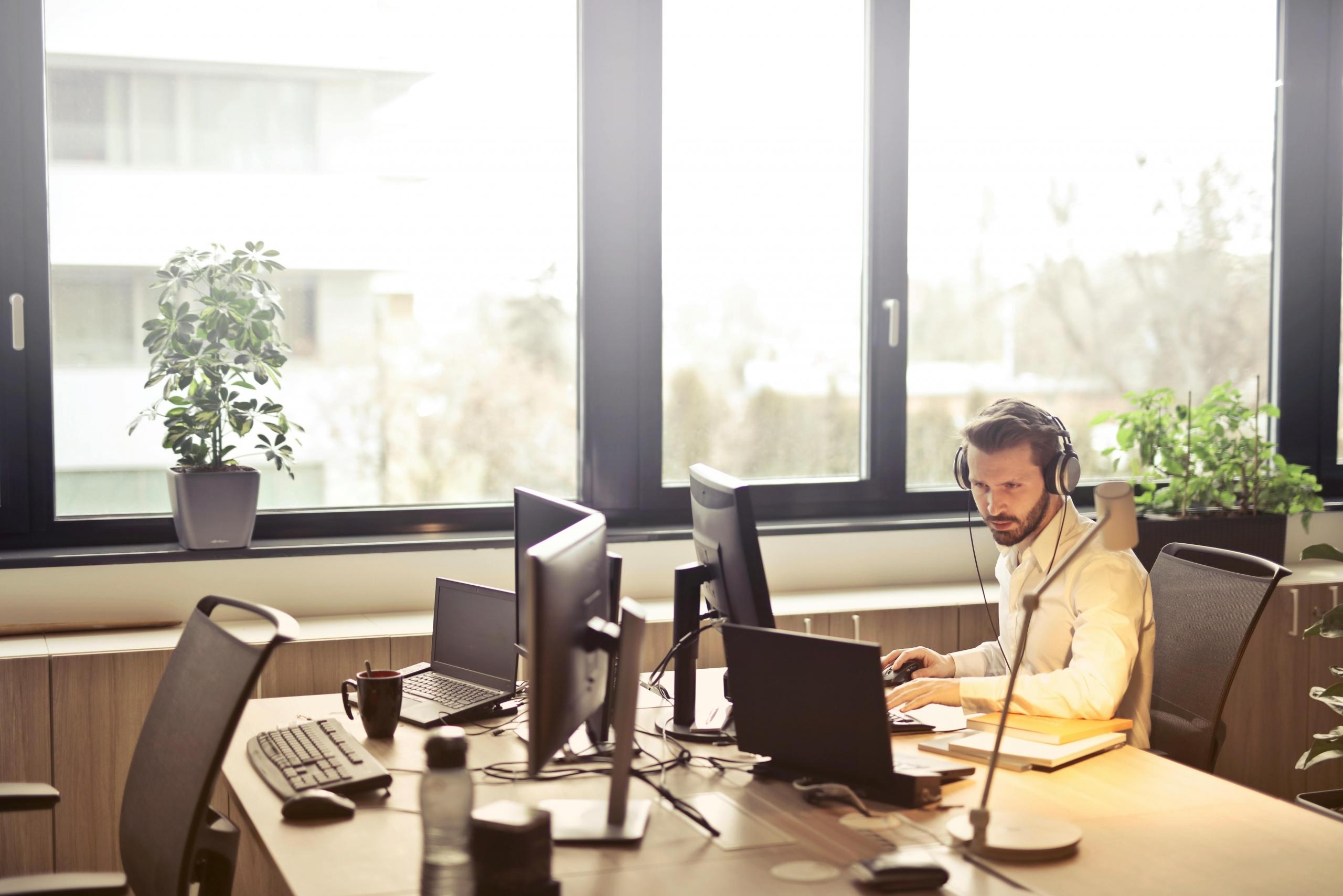 Een man met hoofdtelefoon werkt geconcentreerd aan een bureau met meerdere beeldschermen in een kantoor met grote ramen.