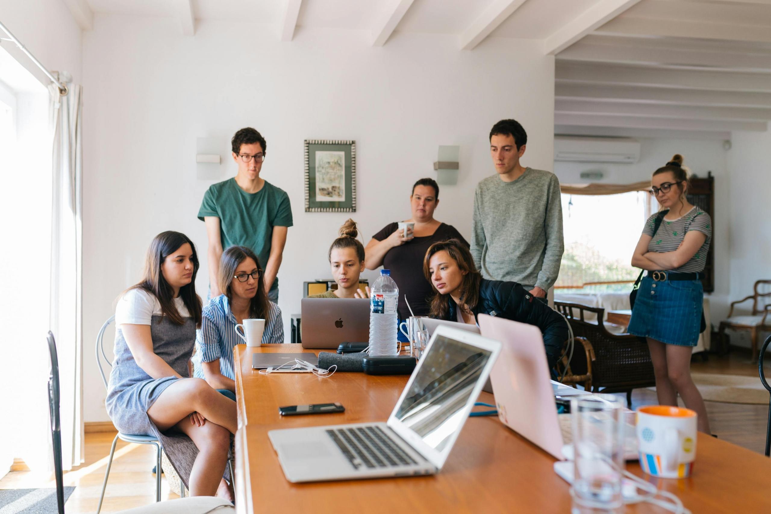 Een groep jonge mensen kijkt naar een laptop op een tafel in een heldere kamer tijdens een informele vergadering of groepsproject.