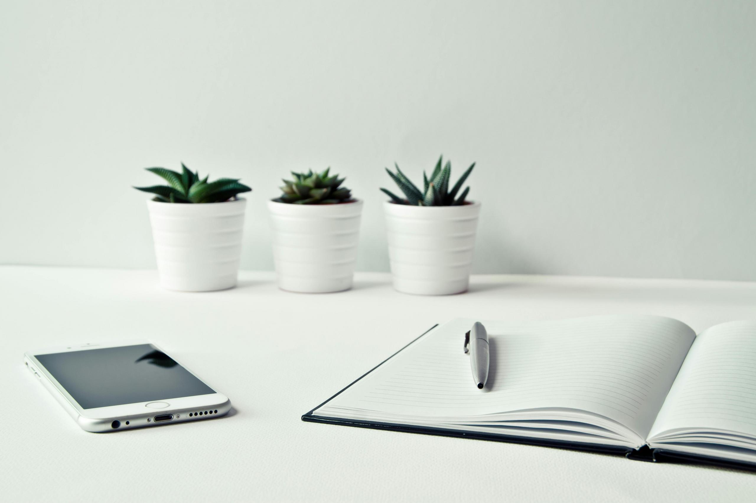 Een eenvoudig, georganiseerd bureau met drie witte bloempotten, een smartphone, en een open notitieboek met een pen erop.