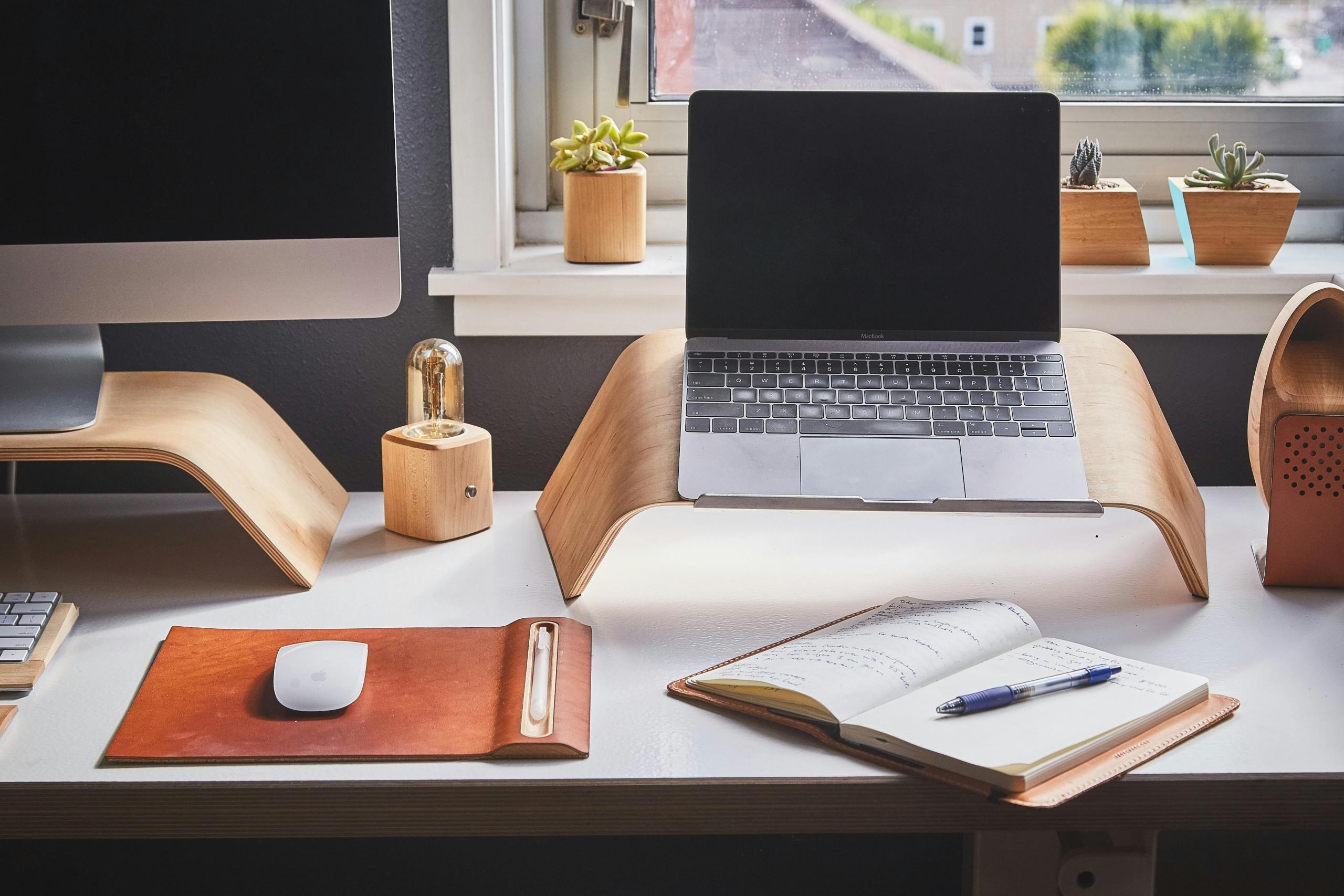 Een georganiseerde werkplek met een iMac, een laptop op een houten standaard, en notitieboekjes omringd door potplanten en stijlvol kantoorbenodigdheden.