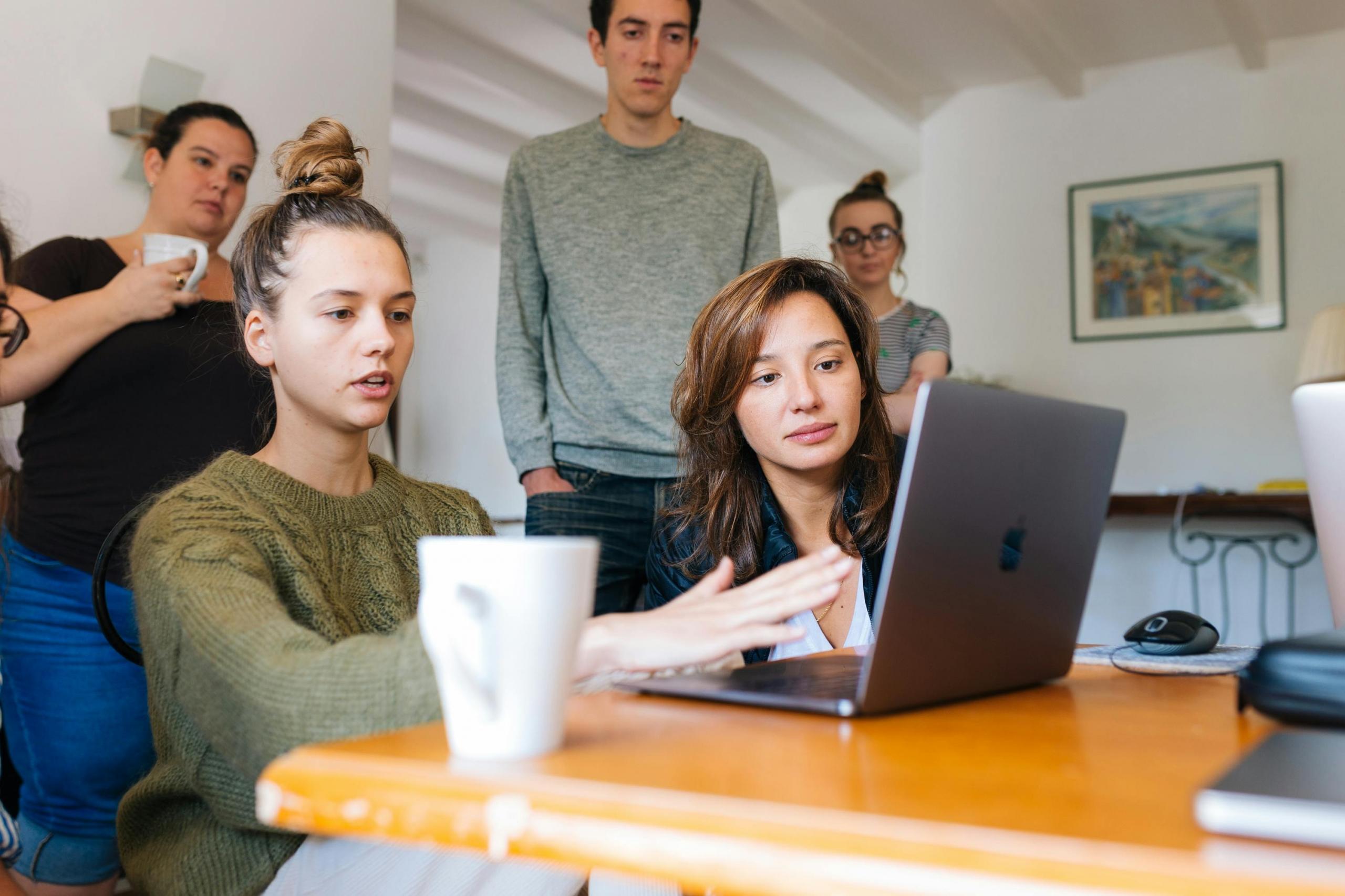 Een groep geconcentreerde jongeren in een informele vergadering rond een tafel met laptops.