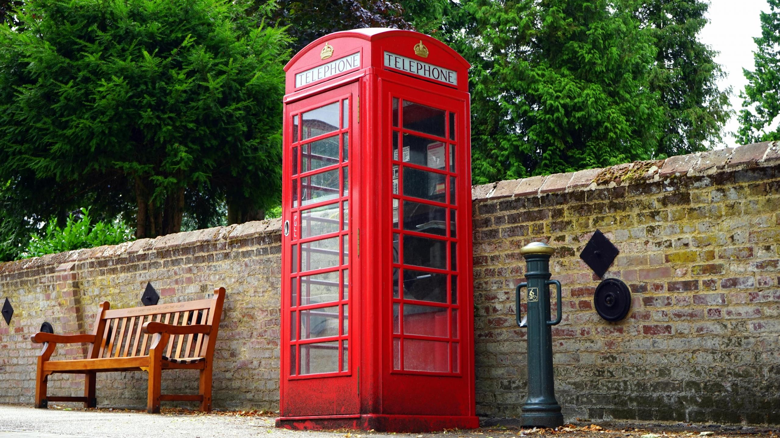 Een klassieke rode Britse telefooncel staat naast een stenen muur met bomen en een houten bankje op de achtergrond.
