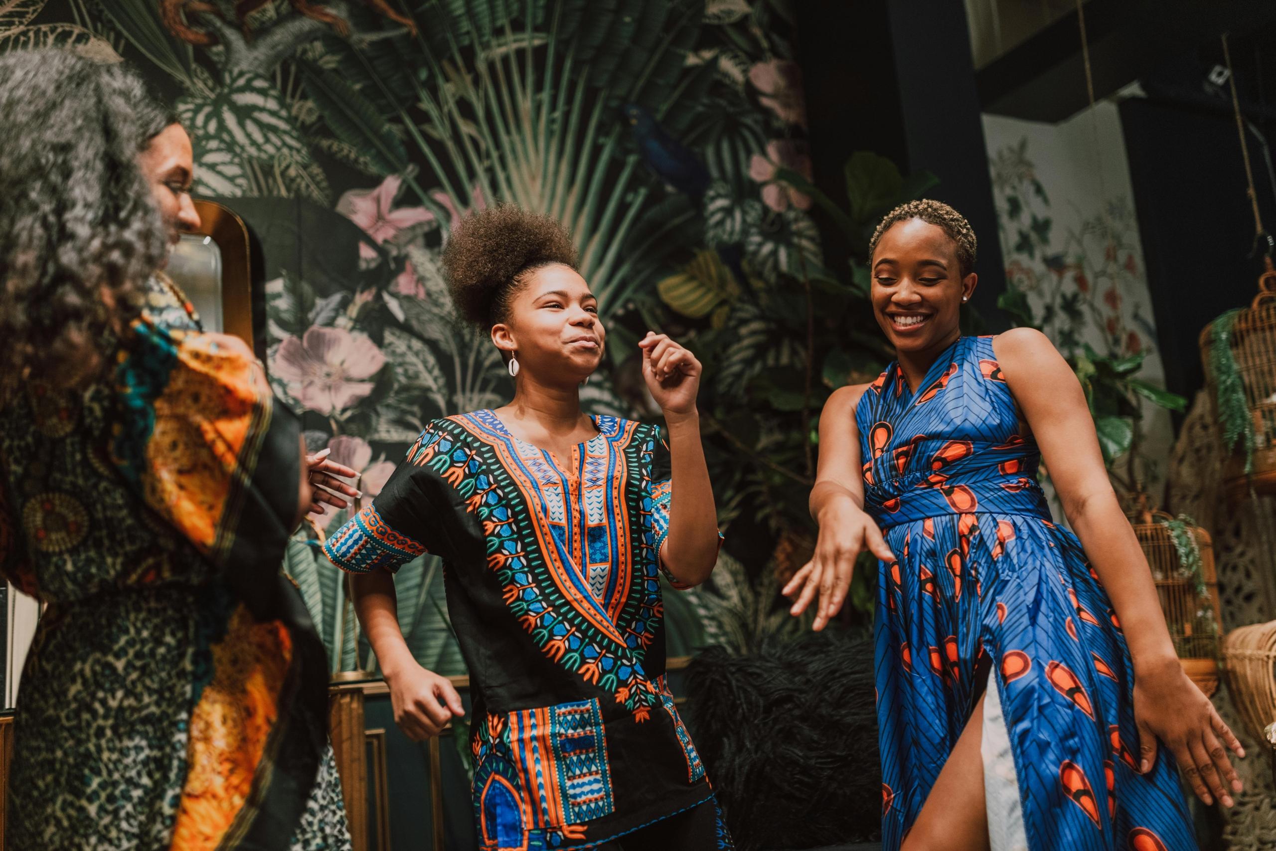 Drie vrouwen in kleurrijke Afrikaanse kleding dansen en lachen samen in een kamer met een exotisch behang.