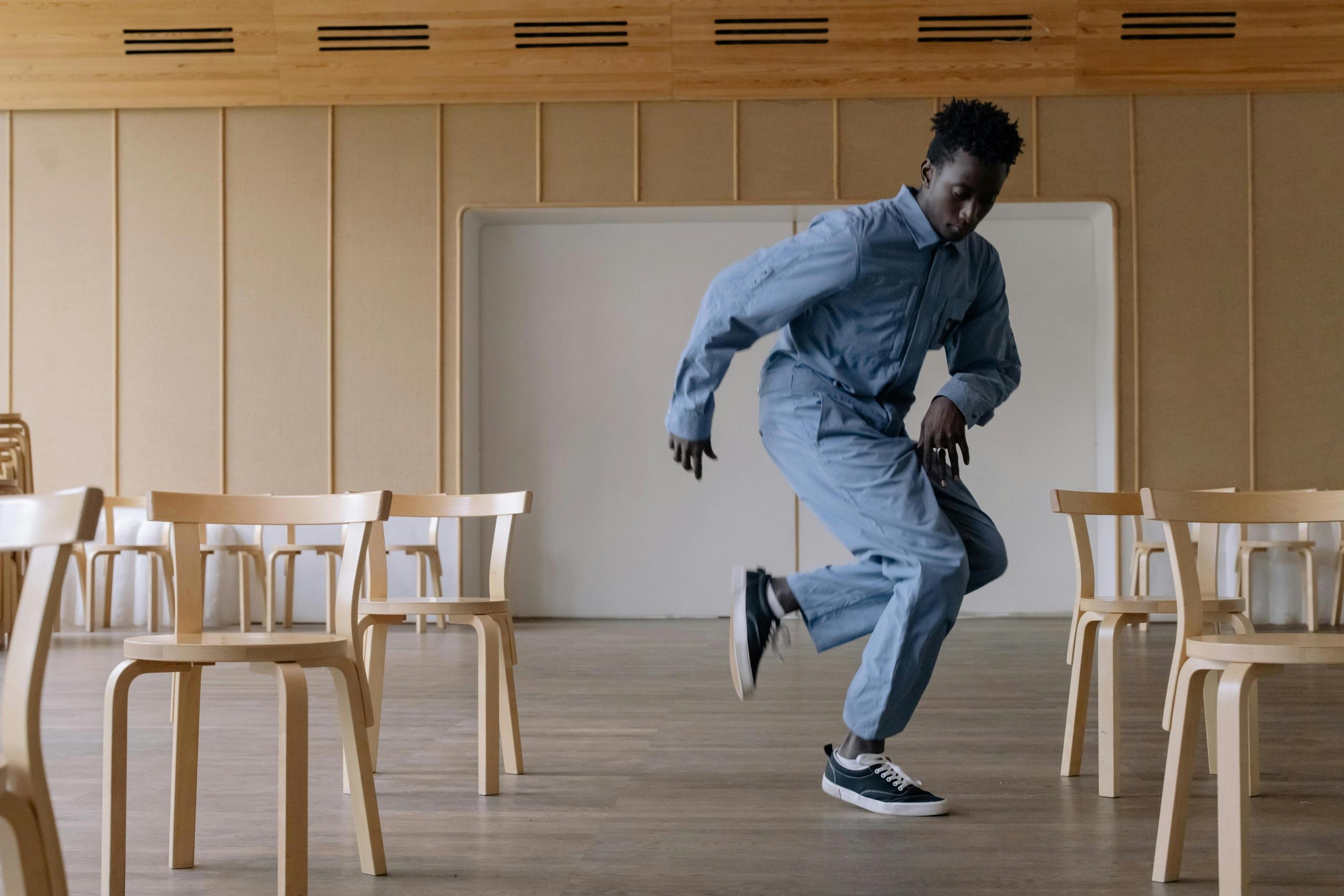 Een man in lichtblauwe kleding danst dynamisch in een zaal met houten stoelen.