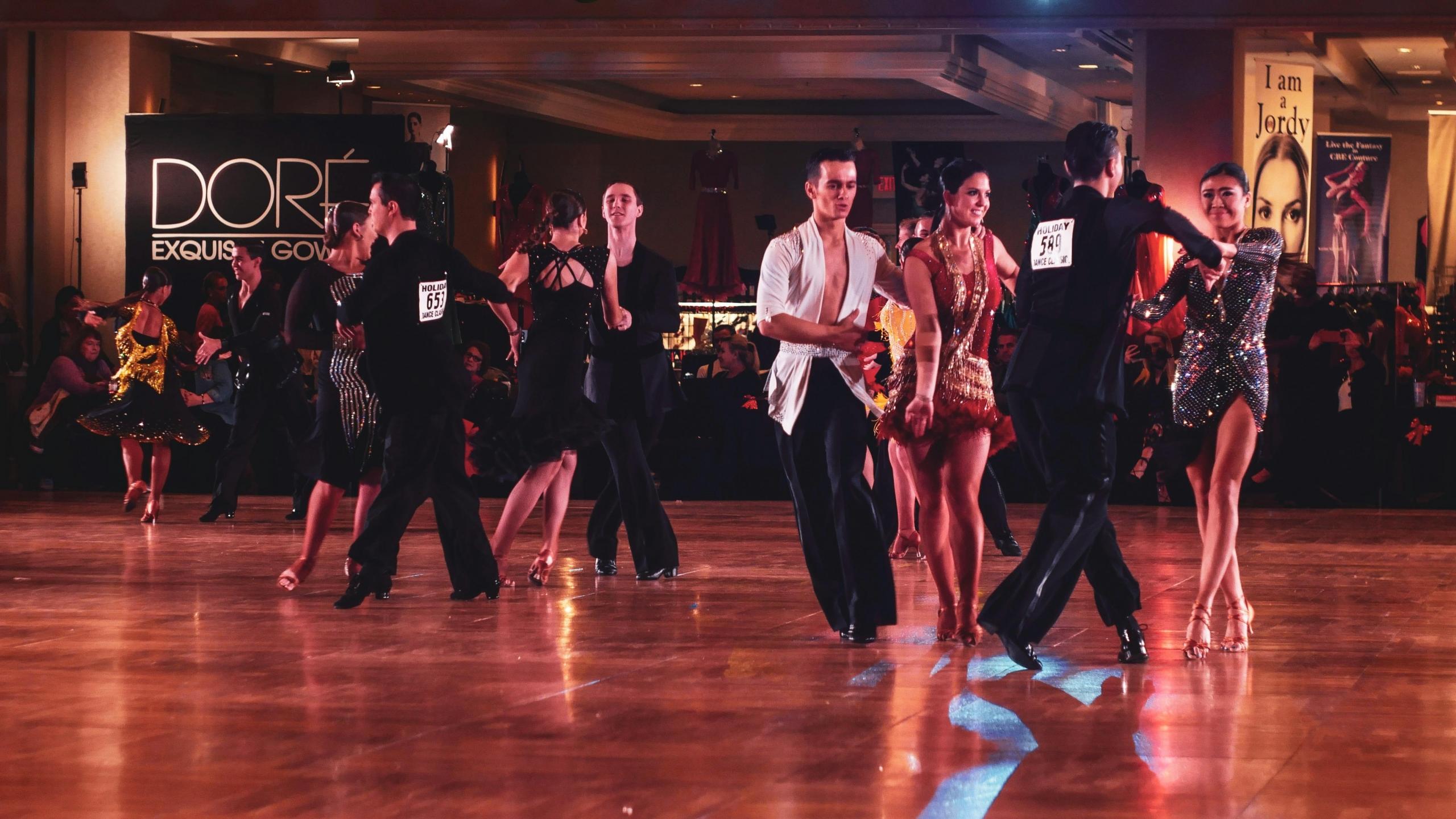 Paren in glinsterende danskostuums voeren ballroomdansen uit op een dansvloer tijdens een competitief evenement.