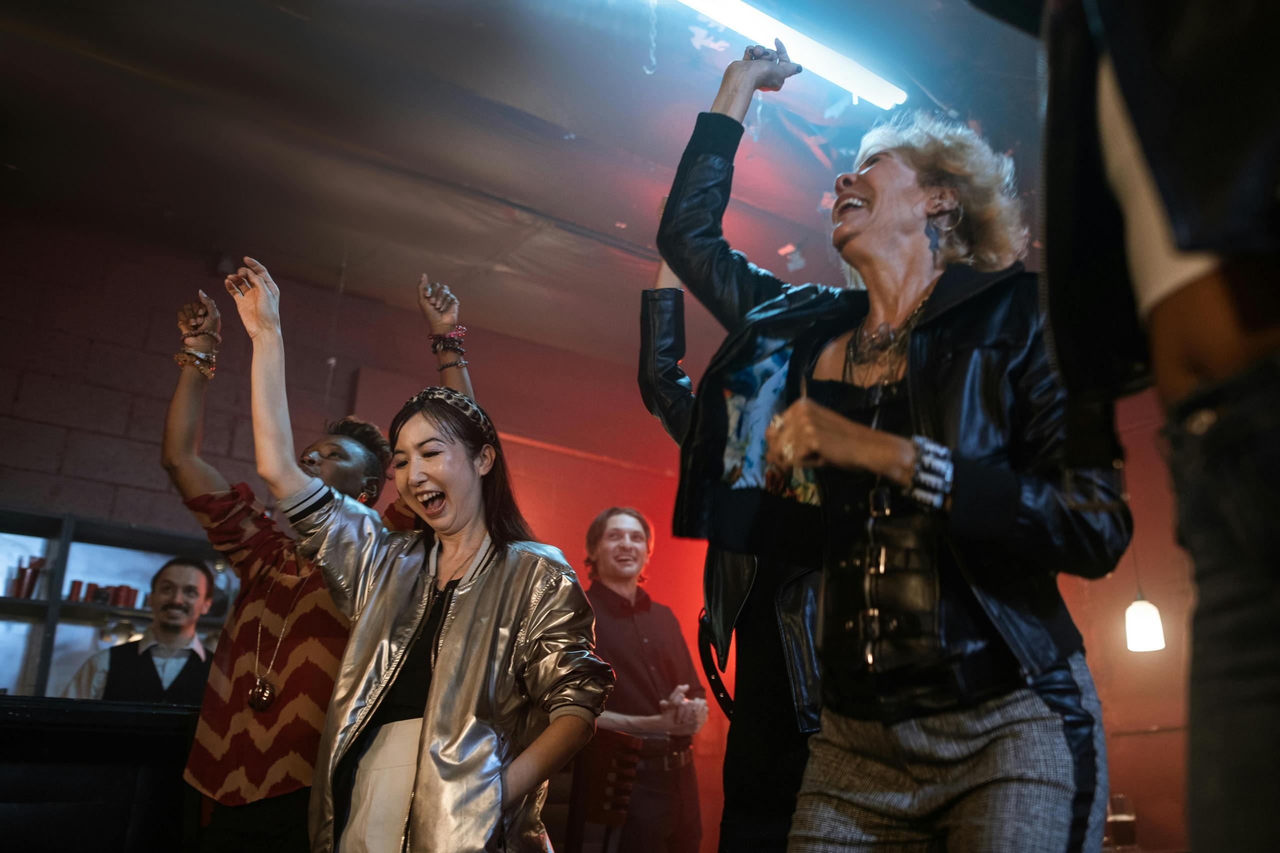 Een gevarieerde groep volwassenen danst en viert feest in een sfeervol verlichte kamer, lachend en met hun armen opgeheven.