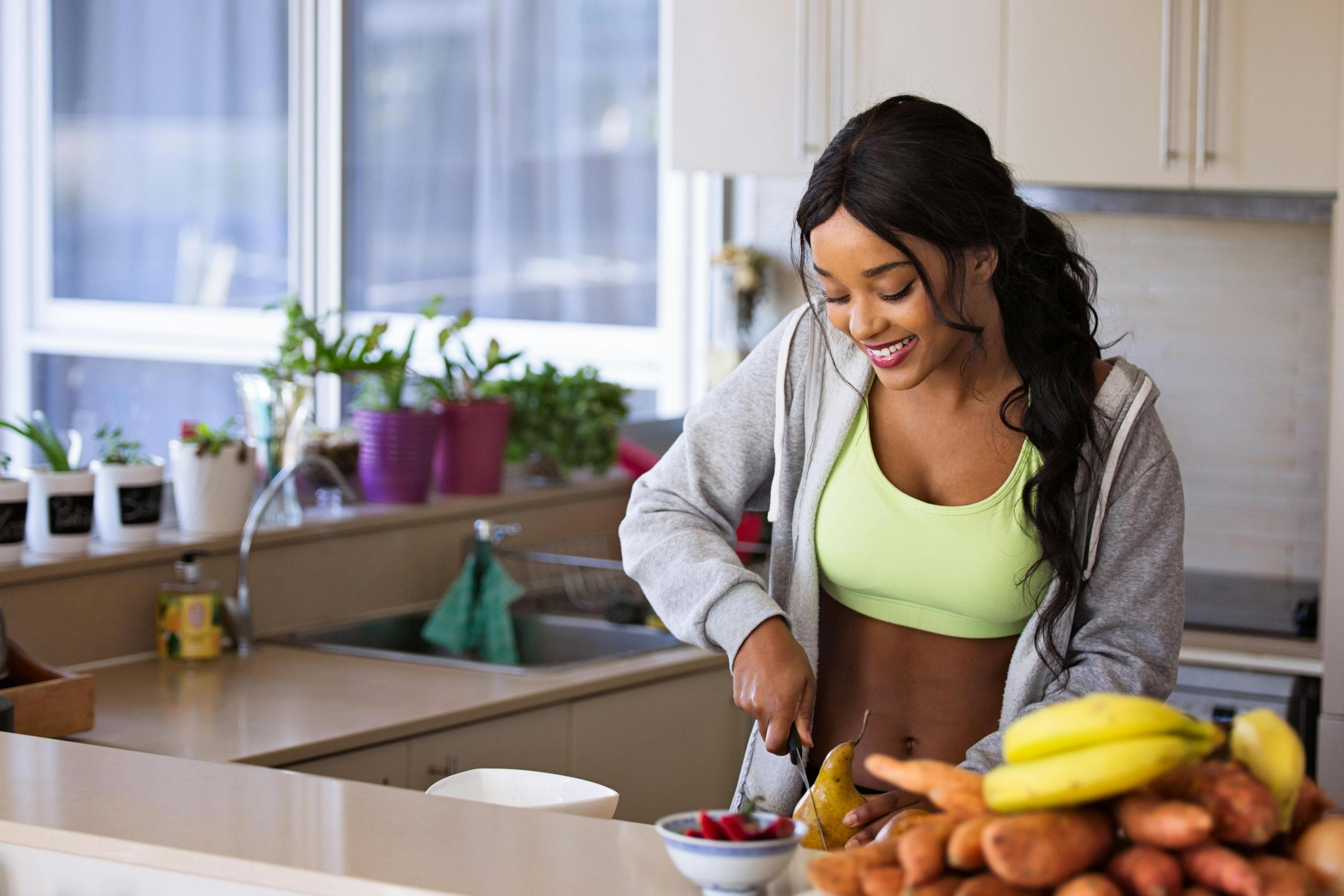 Een vrouw in een lichtgroene sport-bh snijdt fruit in een moderne keuken, glimlachend terwijl ze naar beneden kijkt.