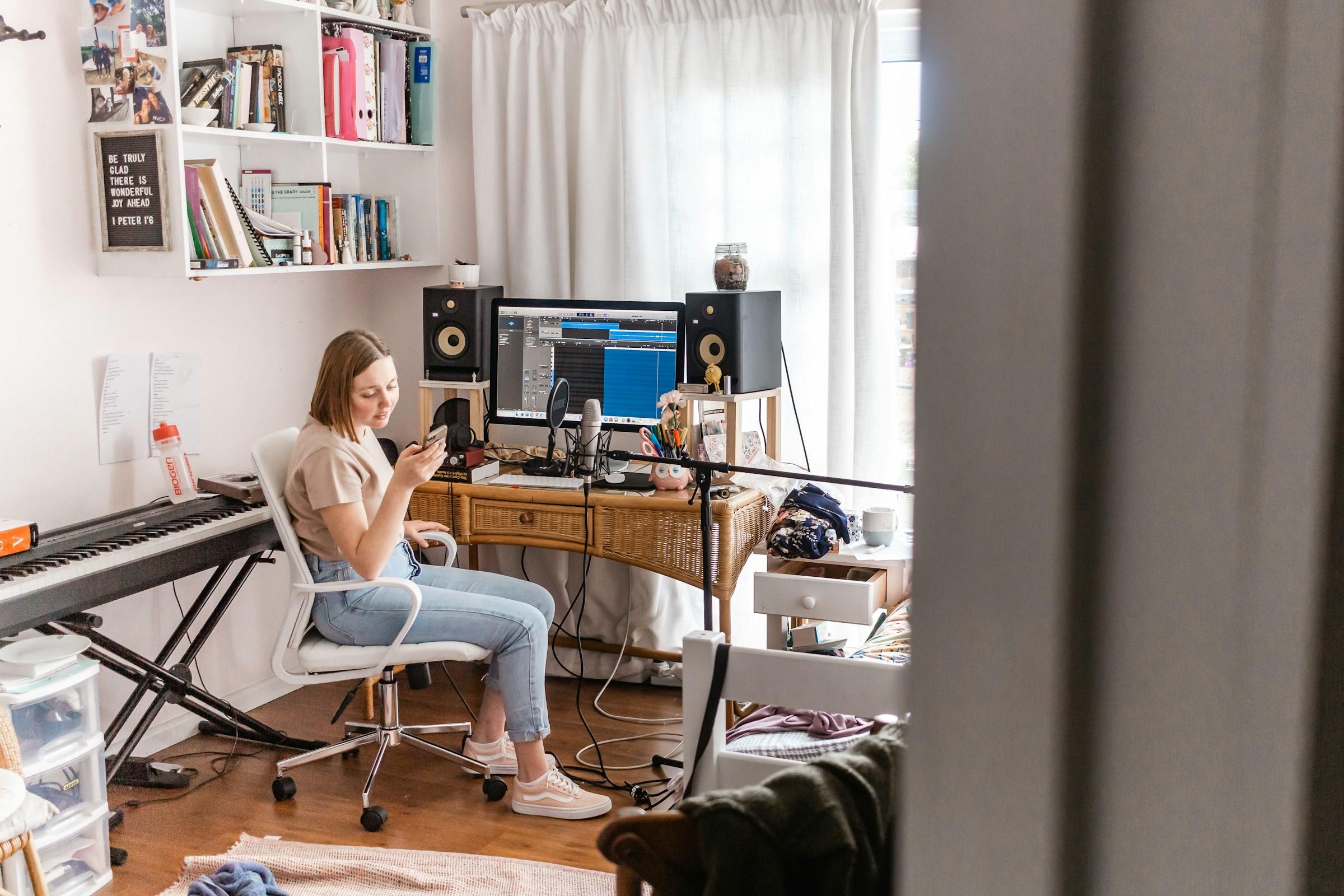 Zittende persoon kijkt op smartphone omringd door muziekapparatuur in kamer