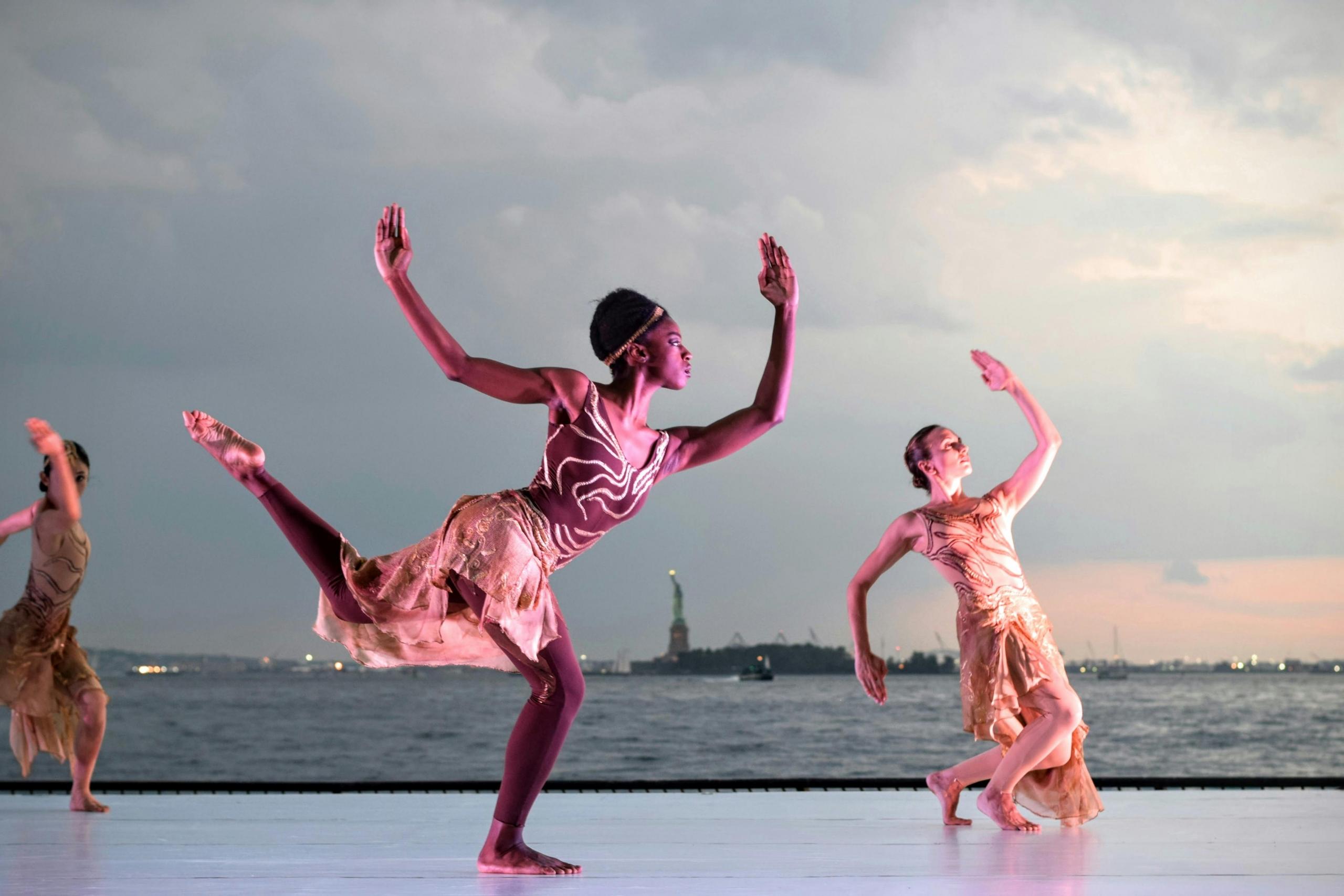 Vrouwelijke dansers in een roze jurk dansen buiten met de skyline op de achtergrond bij zonsondergang.