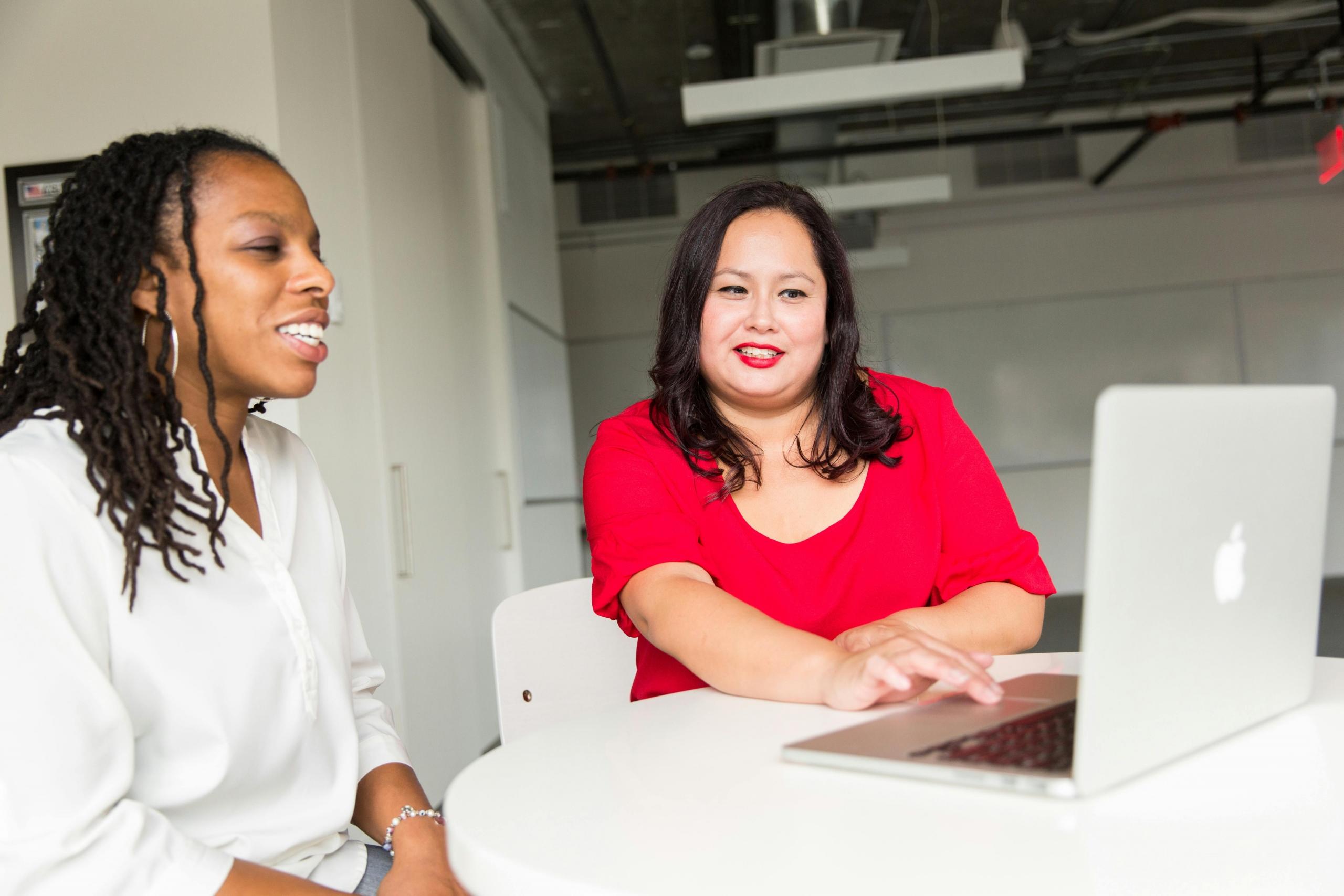 Twee vrouwen in gesprek aan een tafel met een laptop, waarbij een van hen een rode blouse draagt.