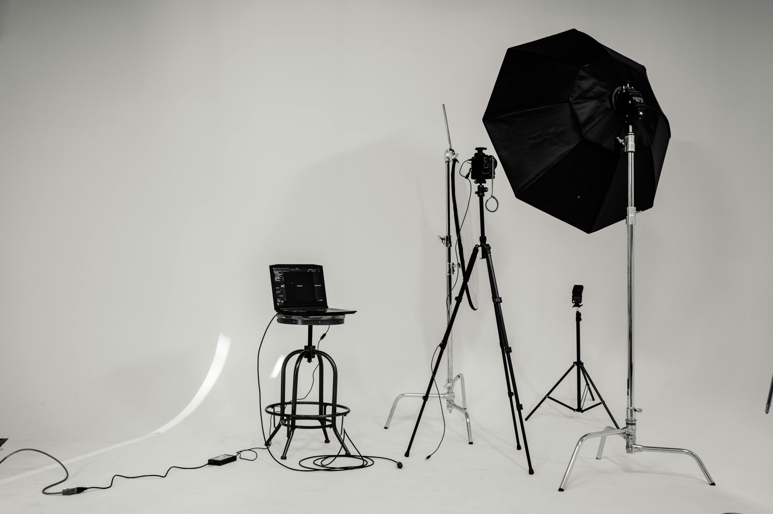 Een professionele fotografiestudio met meerdere lampen, statieven, een camera op een driepoot en een laptop op een standaard tegen een witte achtergrond.