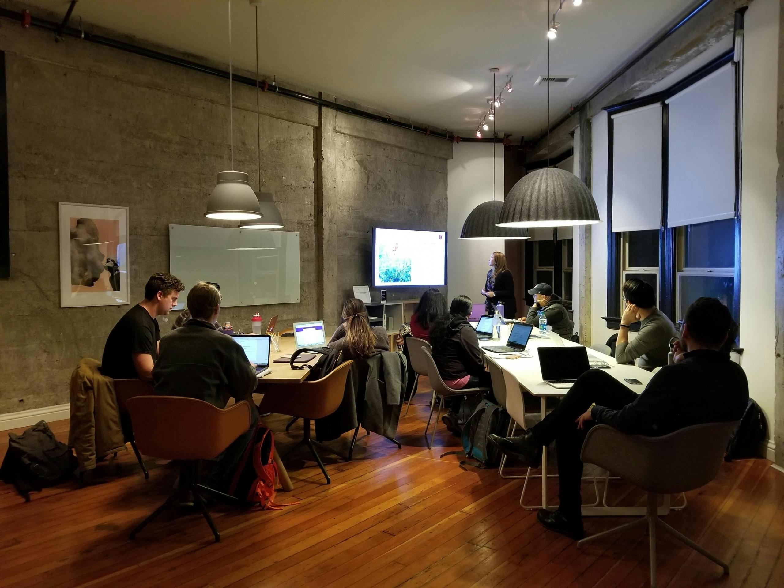 Een groep mensen in een moderne werkruimte luistert naar een presentatie, omringd door computers en sfeerverlichting.