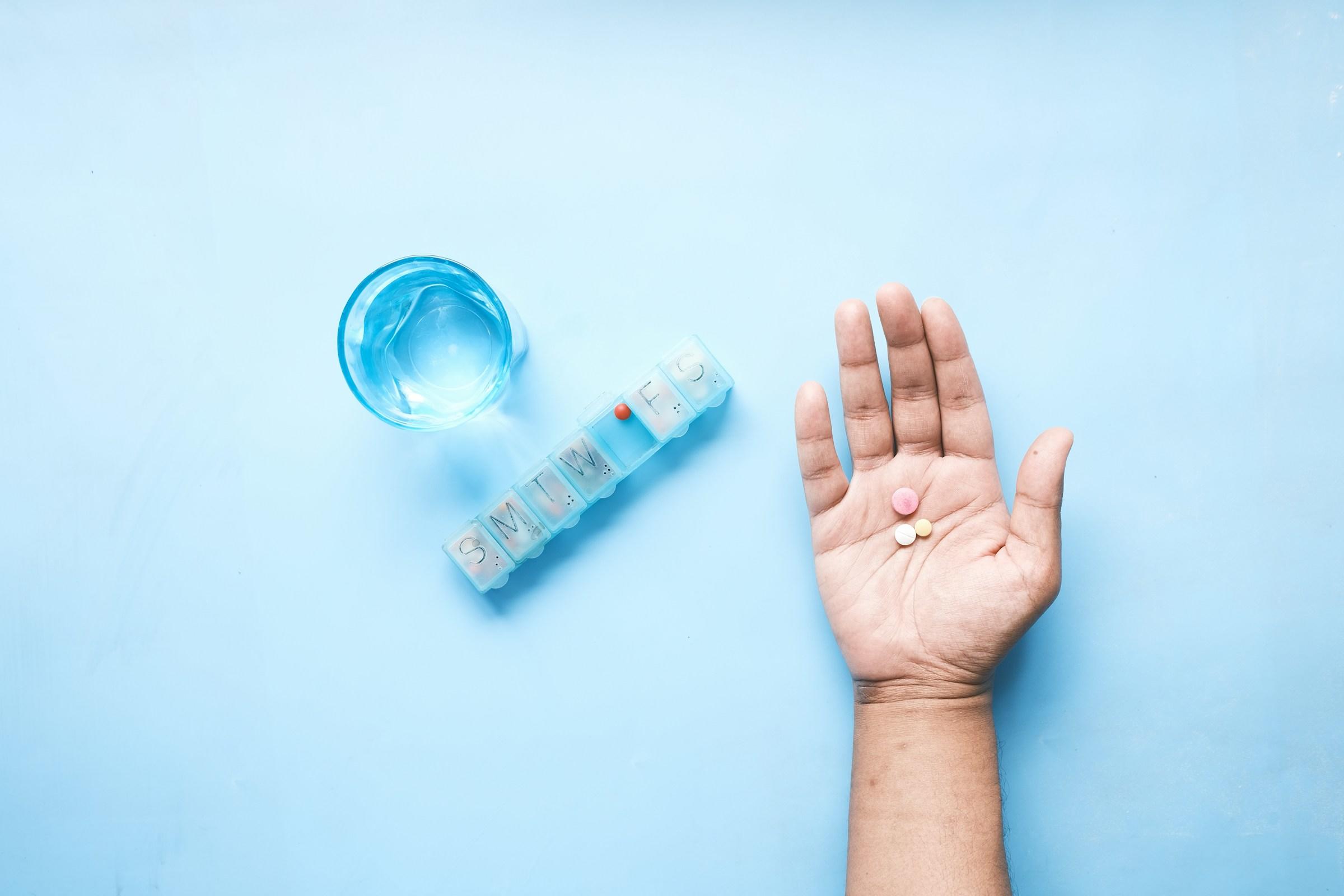 Hand met drie pillen, pillendoosje en glas water op lichtblauwe ondergrond
