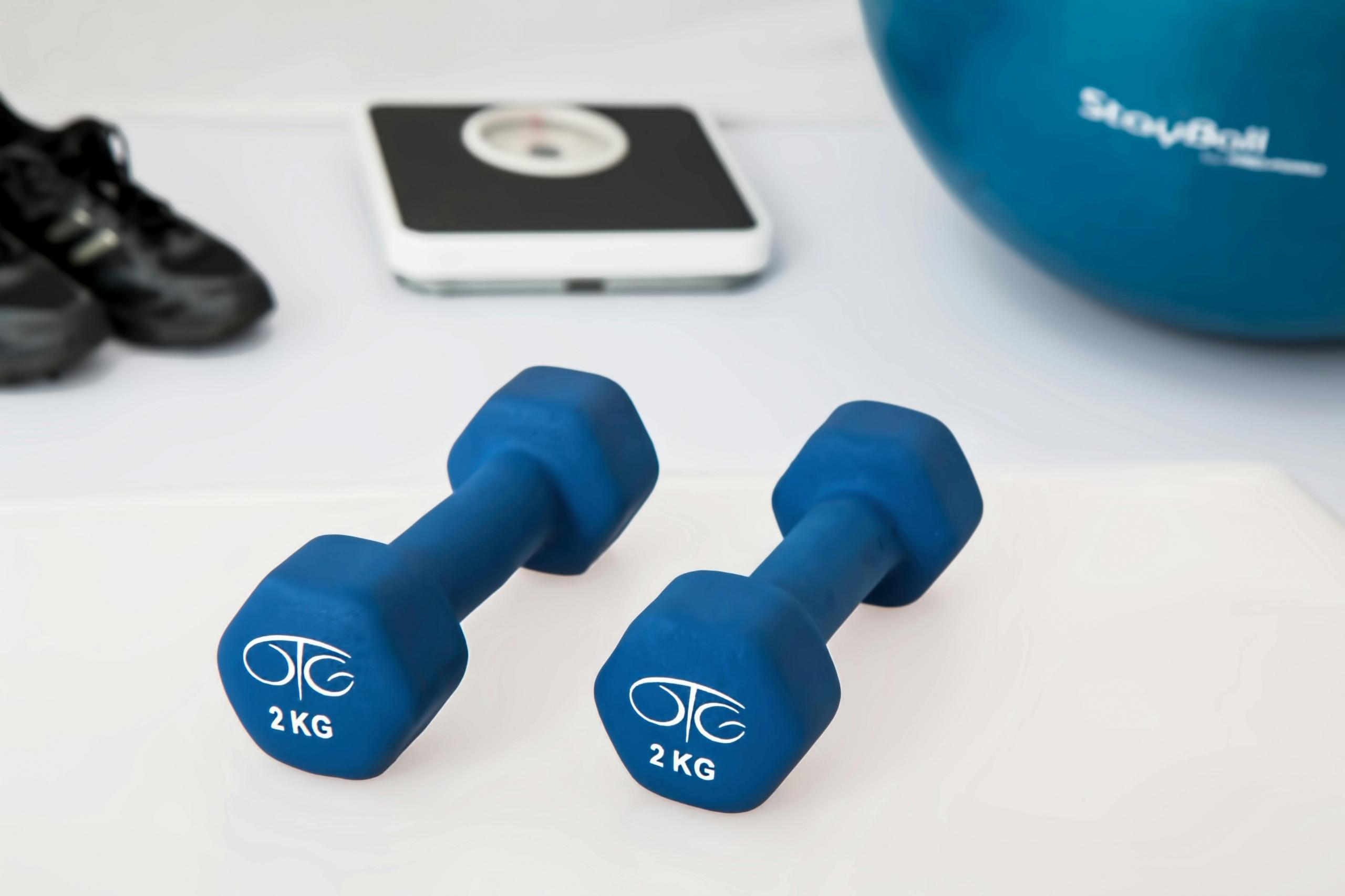 Twee blauwe dumbbells liggen op een witte ondergrond met fitnessapparatuur op de achtergrond.